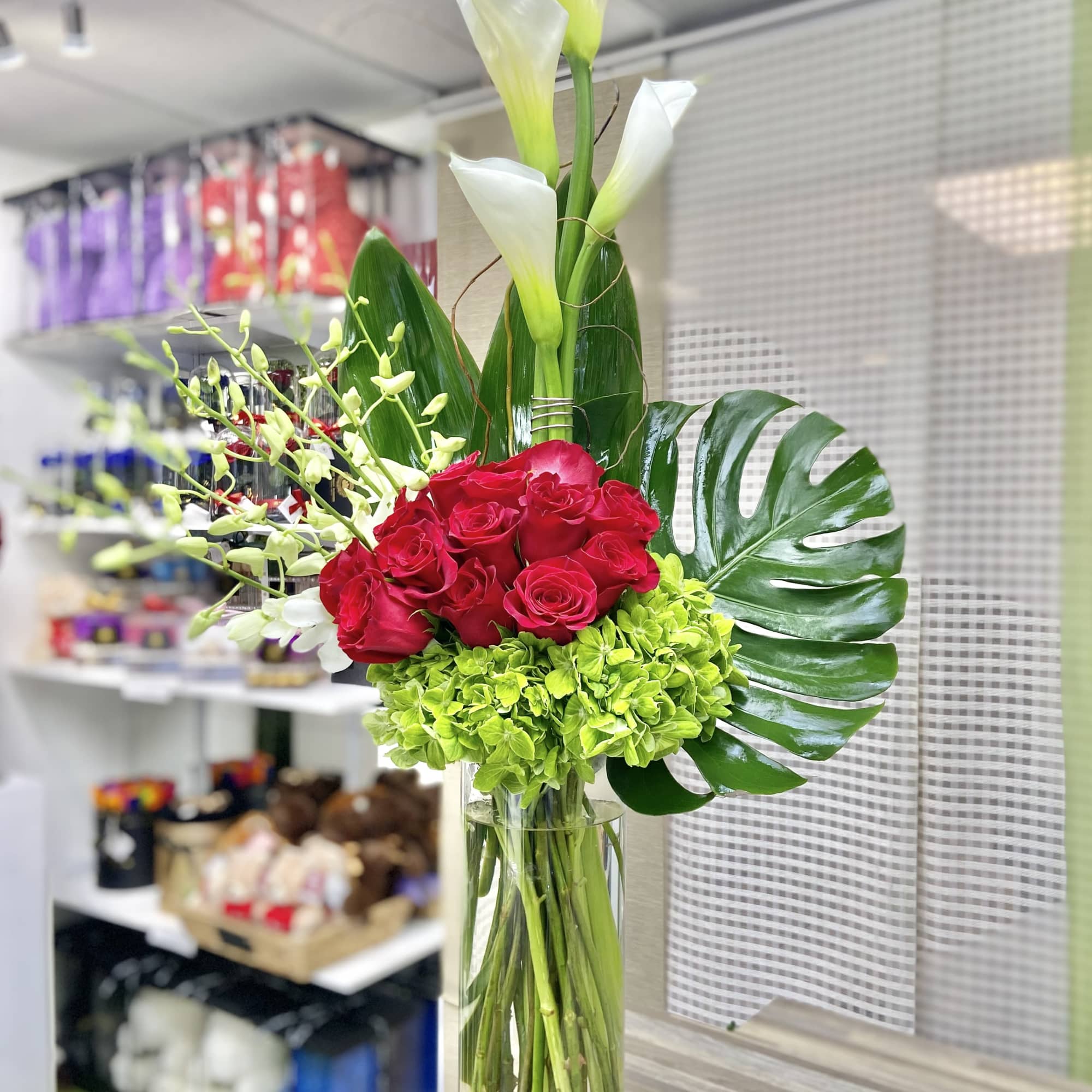 Tall glass vase with white calla lilies, red roses, green hydrangeas, orchids, and large tropical leaves.