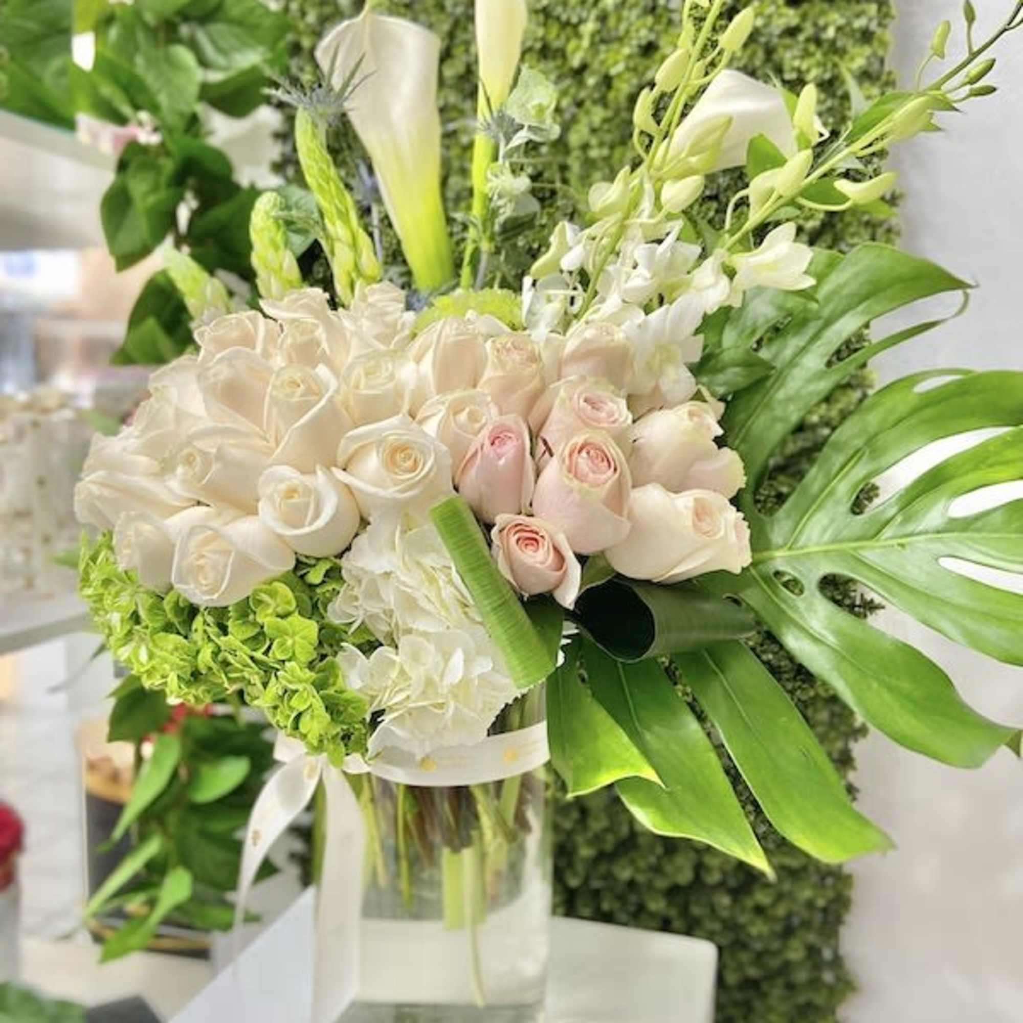 Tall arrangement of white and blush roses, hydrangeas, calla lilies and orchids with large green leaves in a clear glass vase