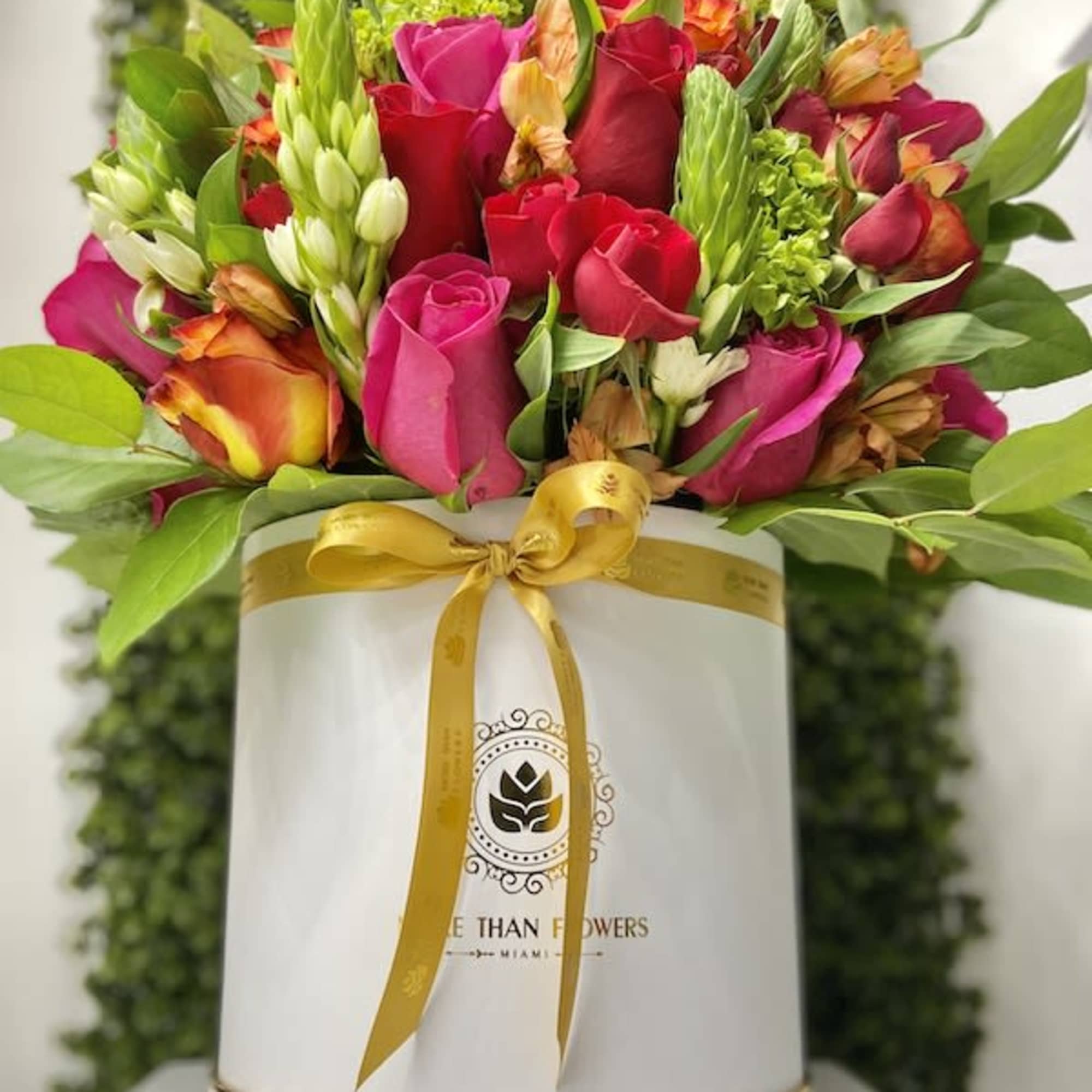 Round white hatbox filled with bright pink and red roses and mixed flowers, tied with a gold ribbon.