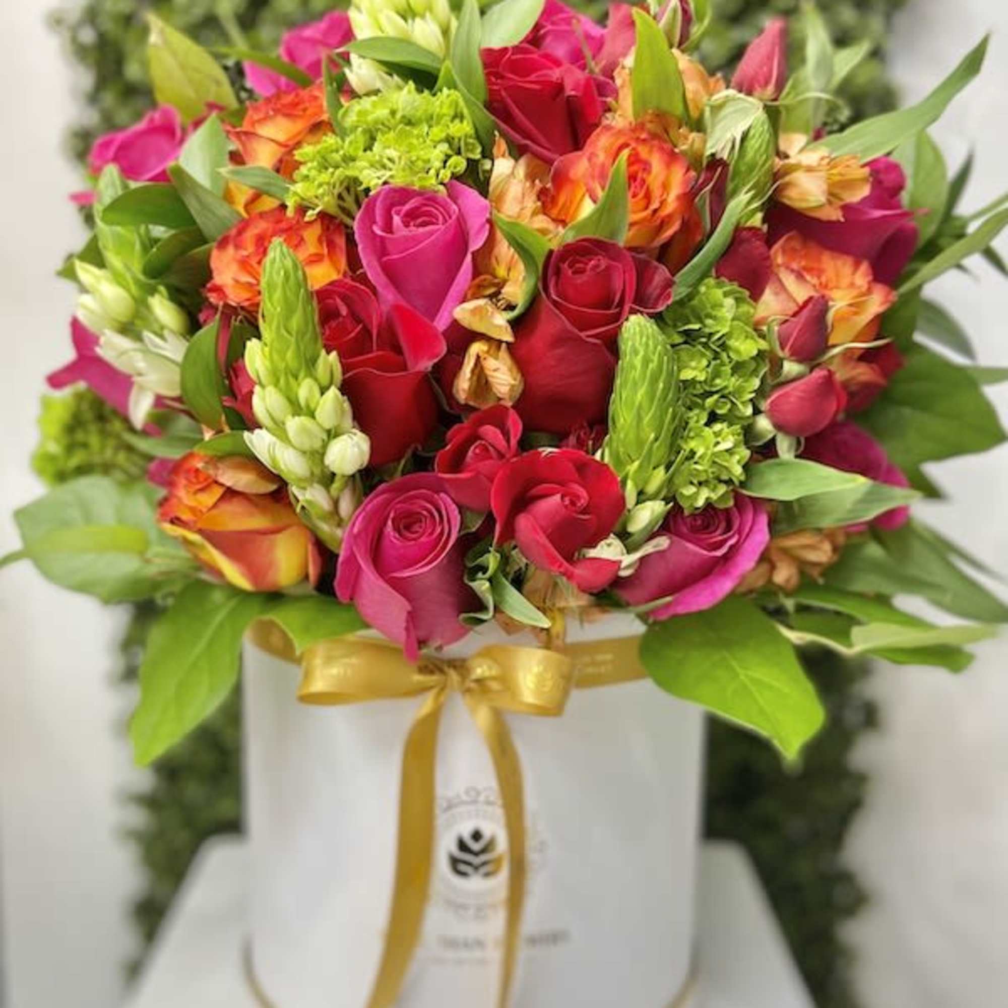 Bright pink, red, and orange flower arrangement in a white hatbox with a gold ribbon.