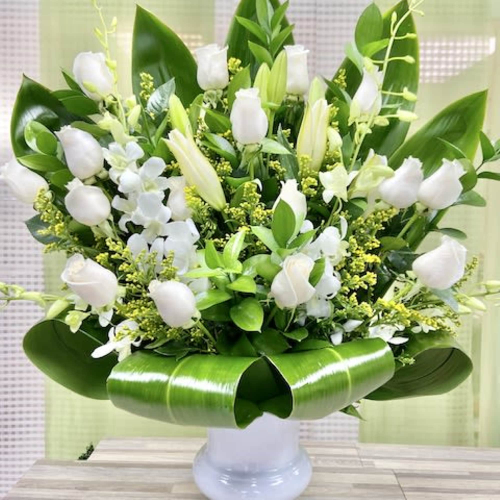 Large arrangement of white roses and lilies with bold looped green leaves in a white pedestal vase