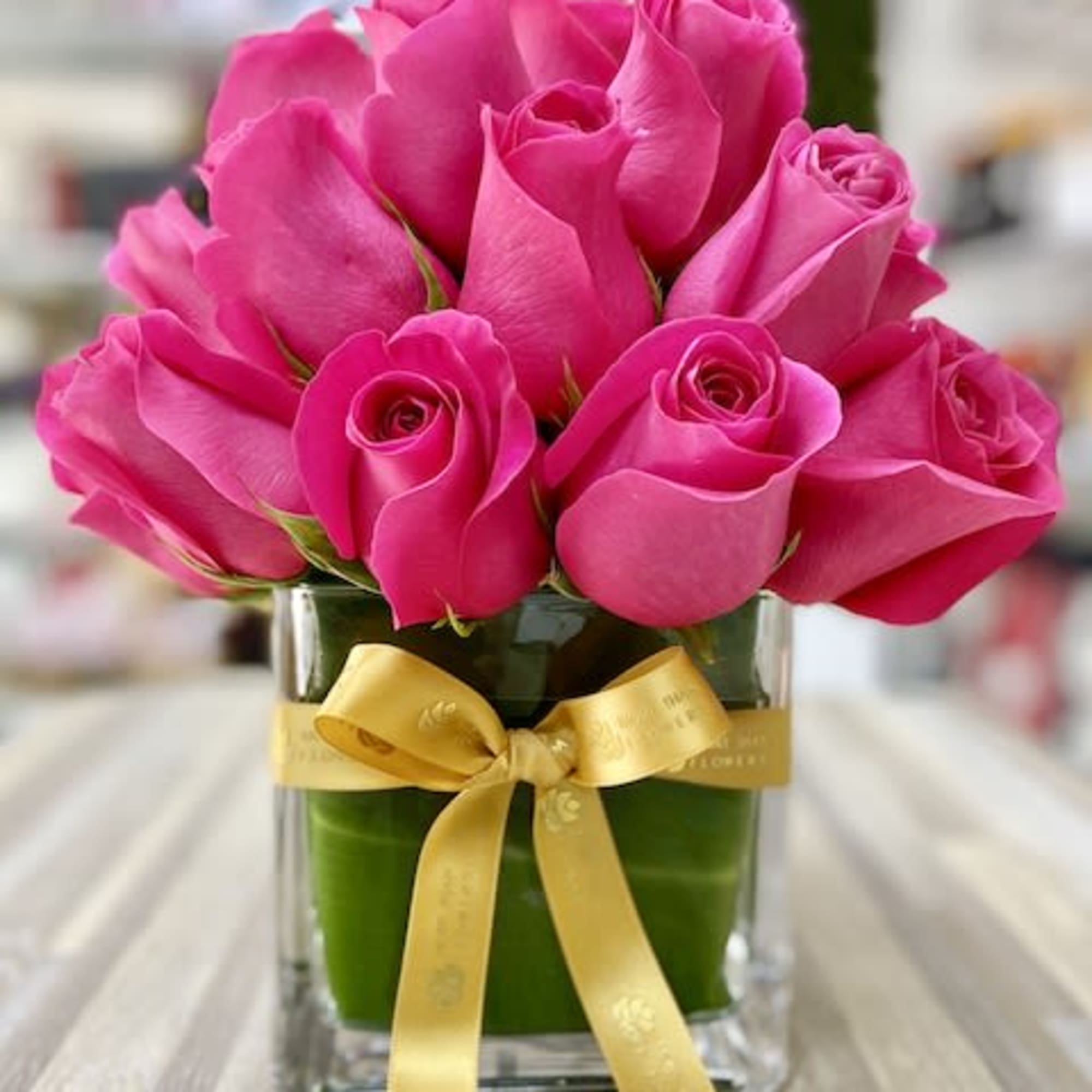 Compact arrangement of hot pink roses in a clear cube vase wrapped with green and tied with a gold ribbon
