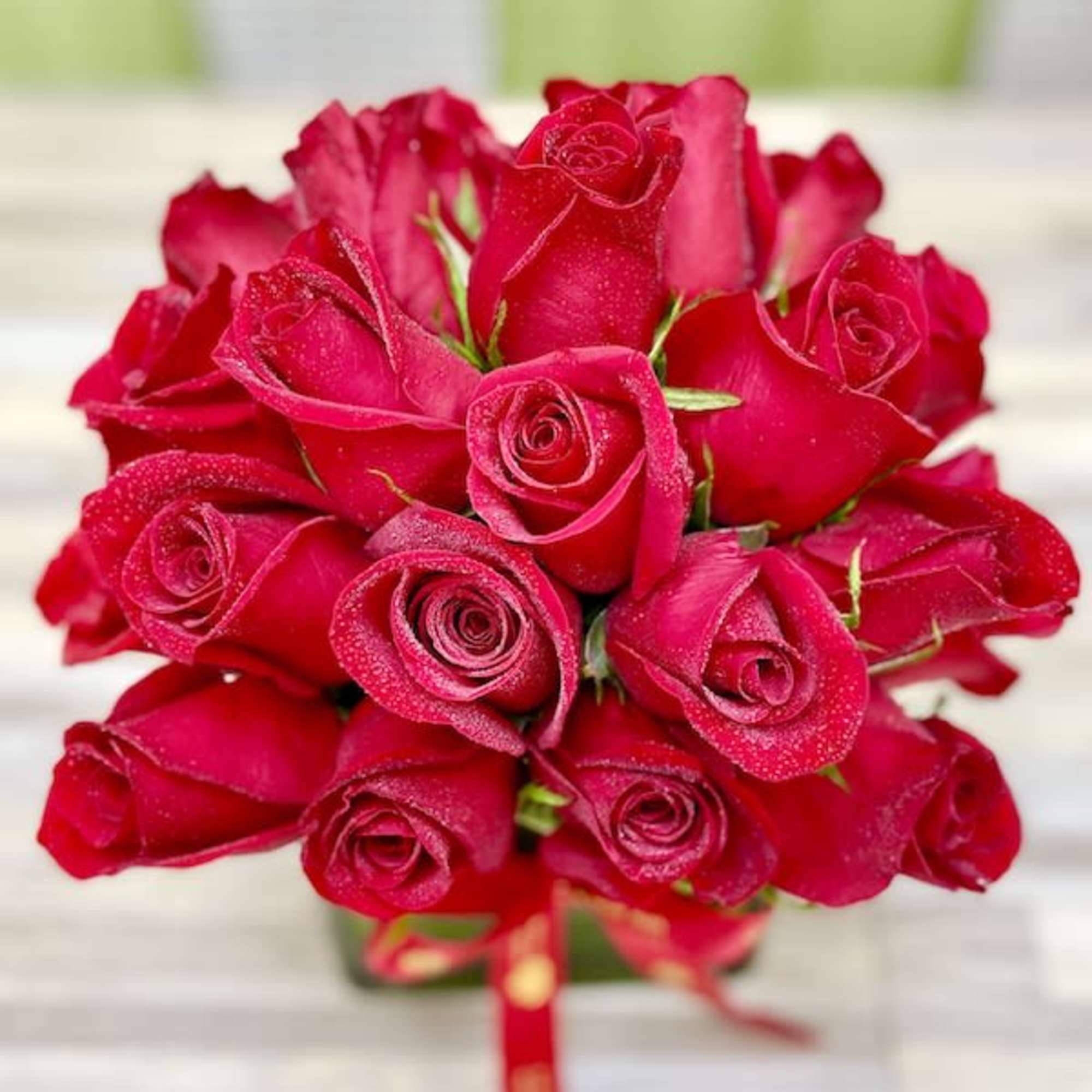 Compact bouquet of red roses with dewdrops in a square container with a red ribbon.