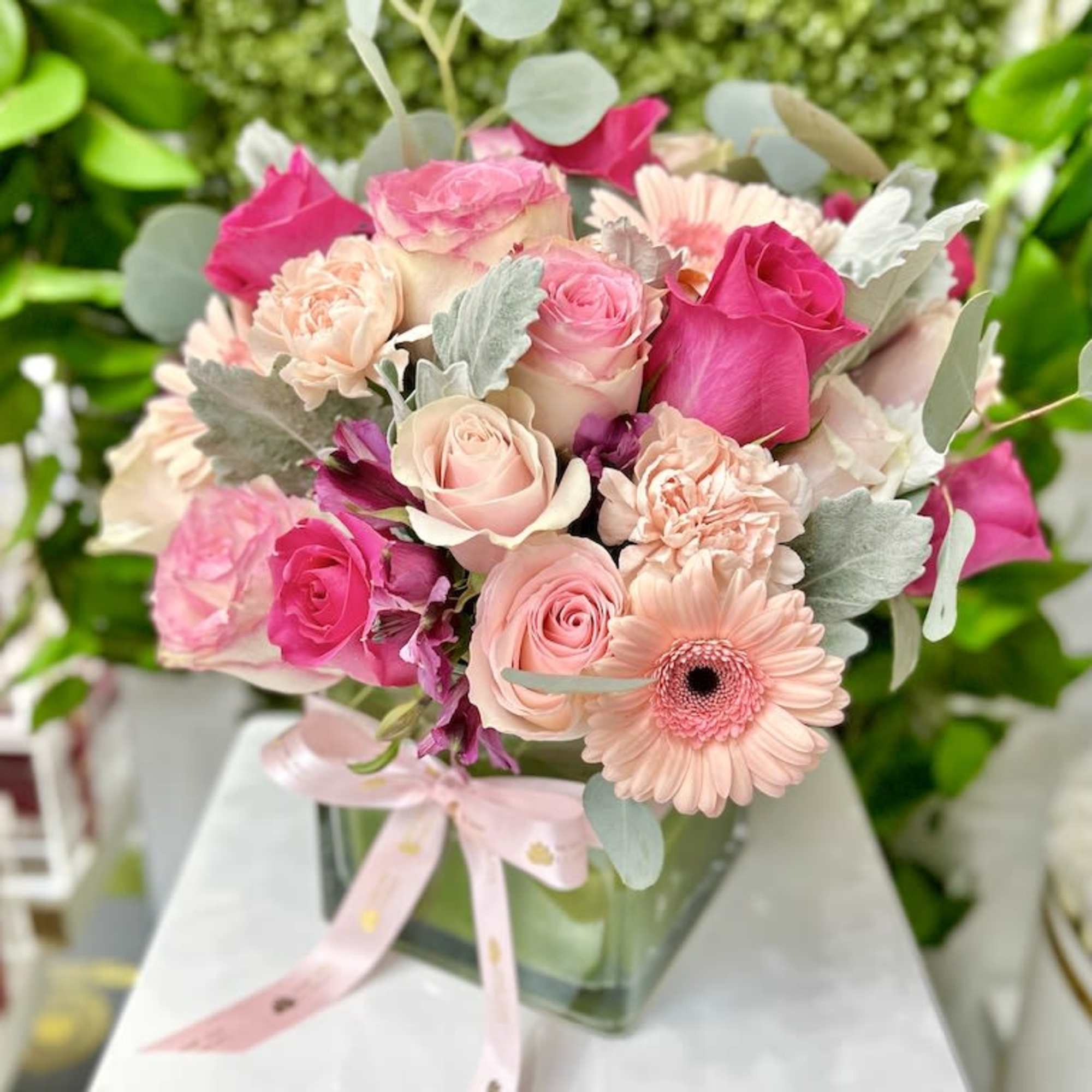 Pink and peach roses, carnations, and gerbera daisies in a glass cube vase with a pink ribbon bow