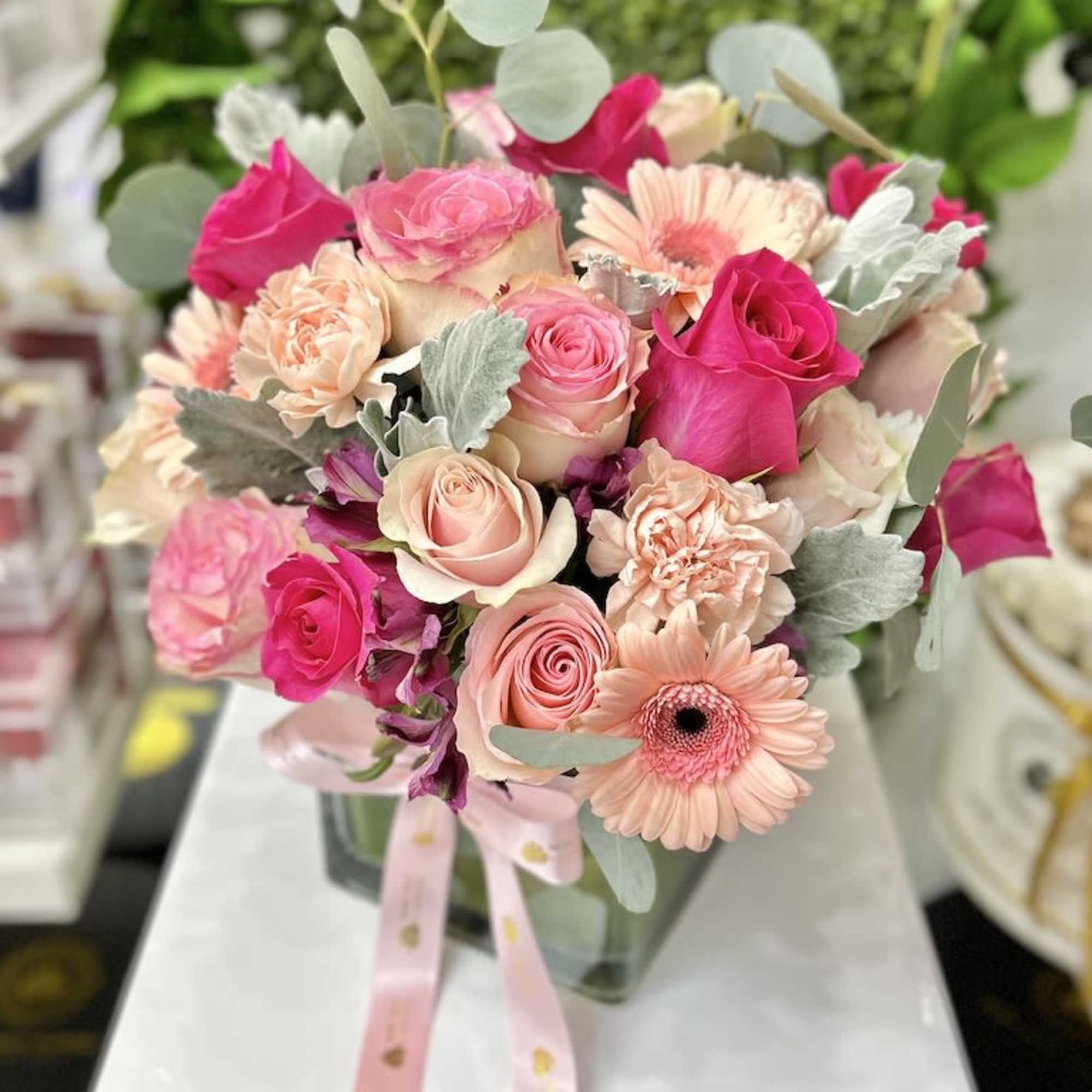 Low arrangement of hot pink and blush roses, gerbera daisies, and carnations in a glass cube vase with pink ribbon