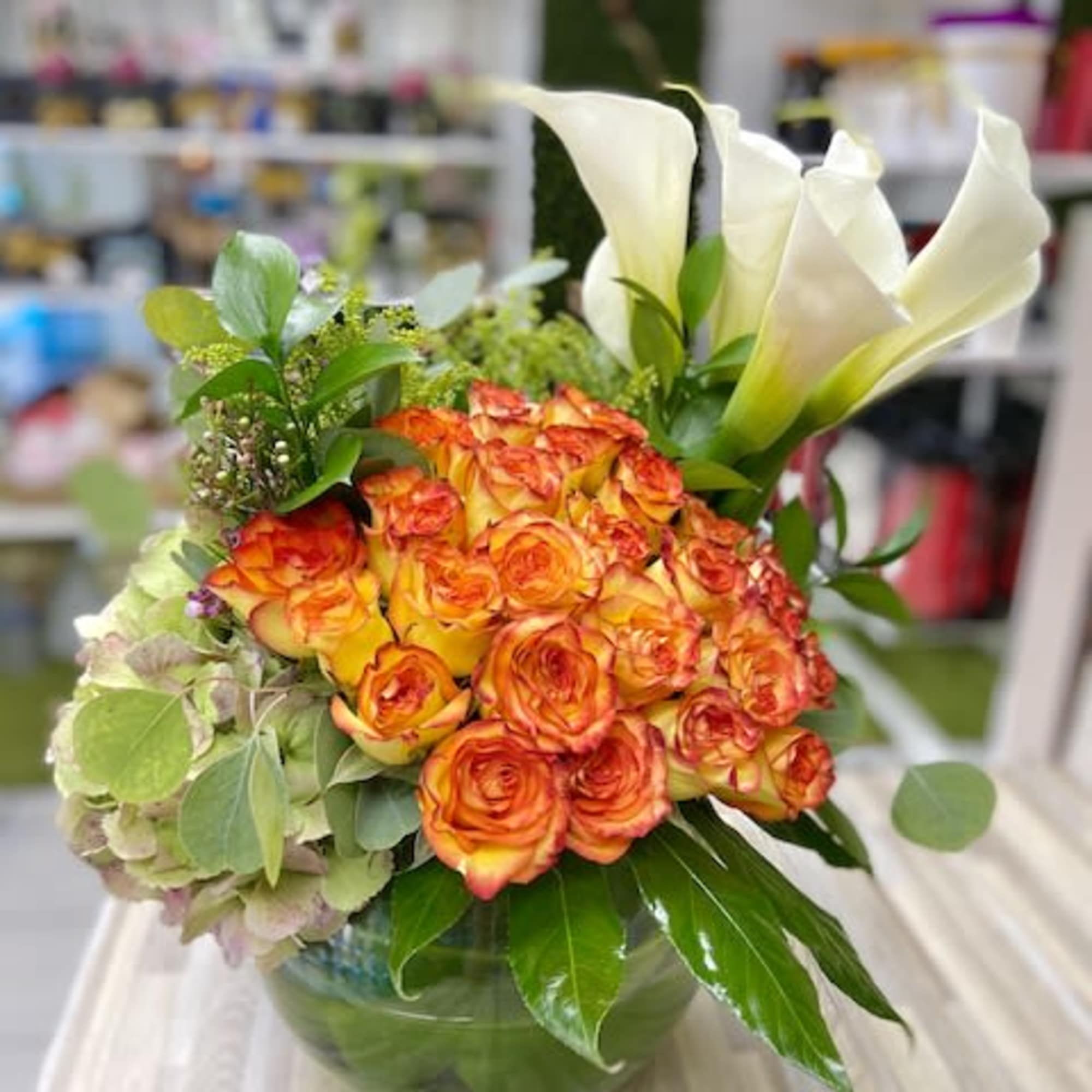Low glass bowl arrangement of orange roses, white calla lilies, and hydrangea.