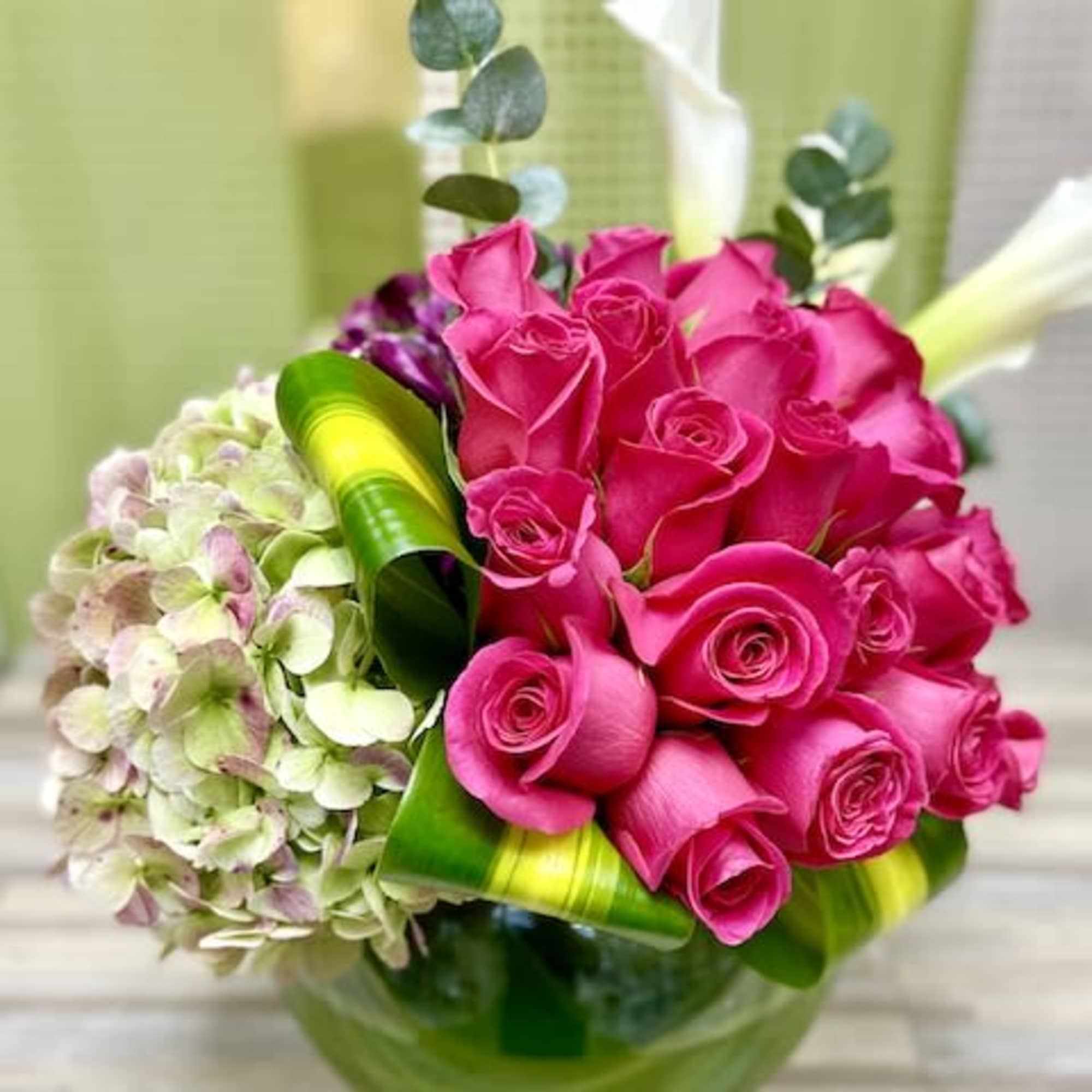 Compact arrangement of hot pink roses, green hydrangea, and white calla lilies in a round green glass vase