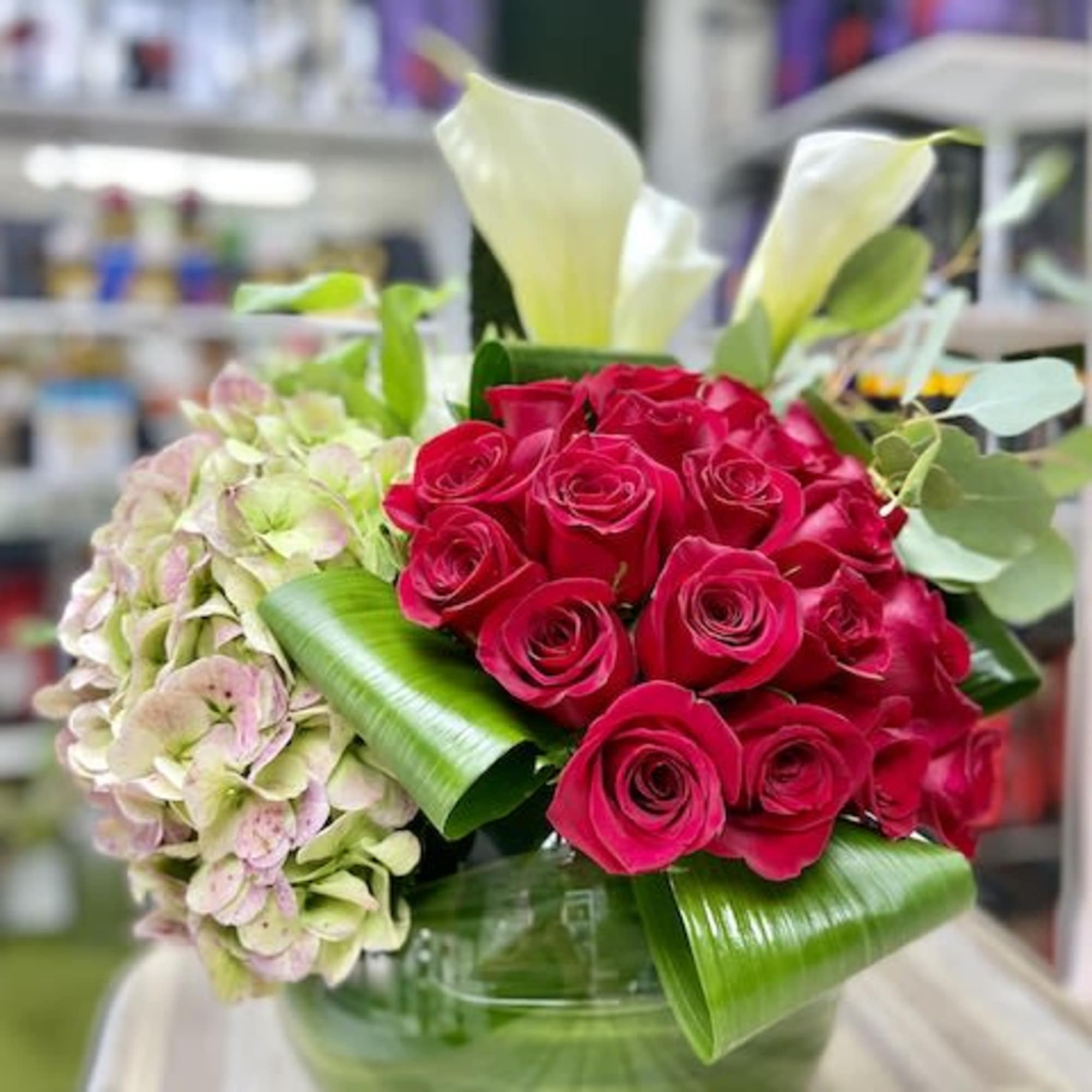 Low glass vase arrangement with red roses, white calla lilies and hydrangea with folded green leaves.