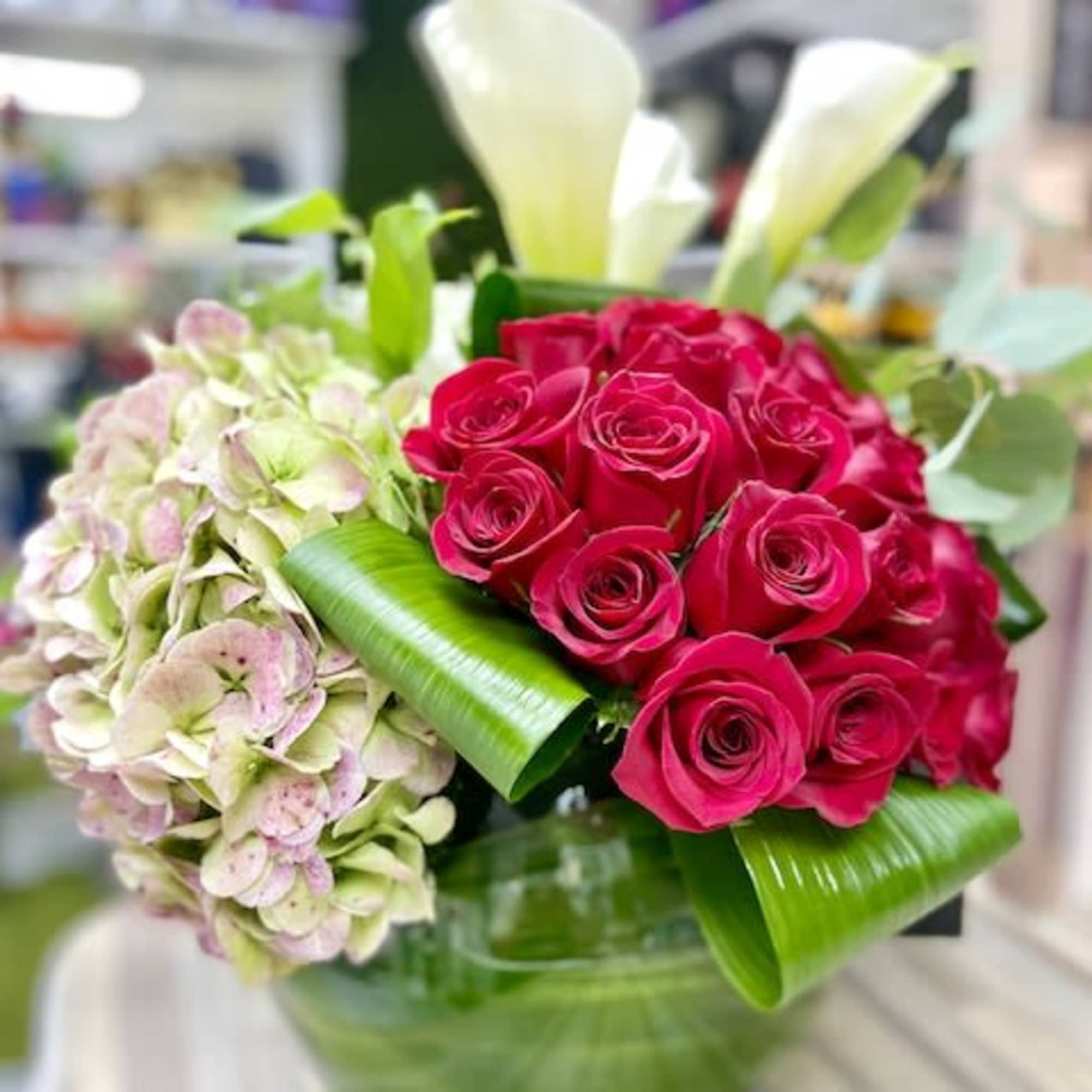 Low glass vase arrangement with hot pink roses, pale hydrangea, white calla lilies, and folded green leaves.