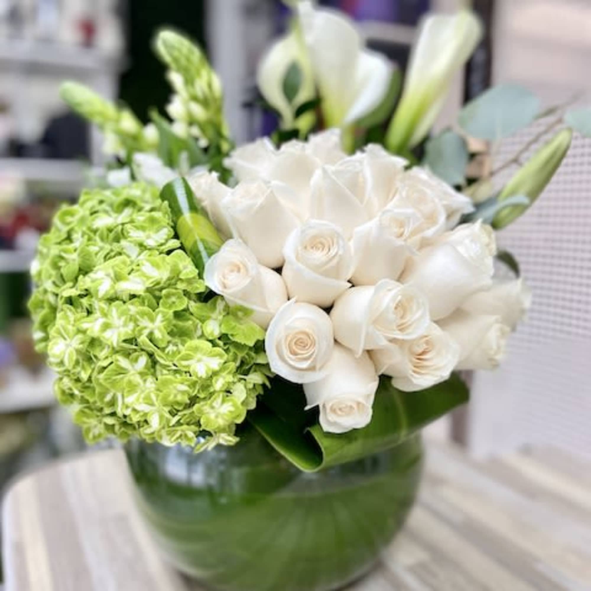 Low round arrangement of white roses, white calla lilies, and green hydrangeas in a clear glass bowl.