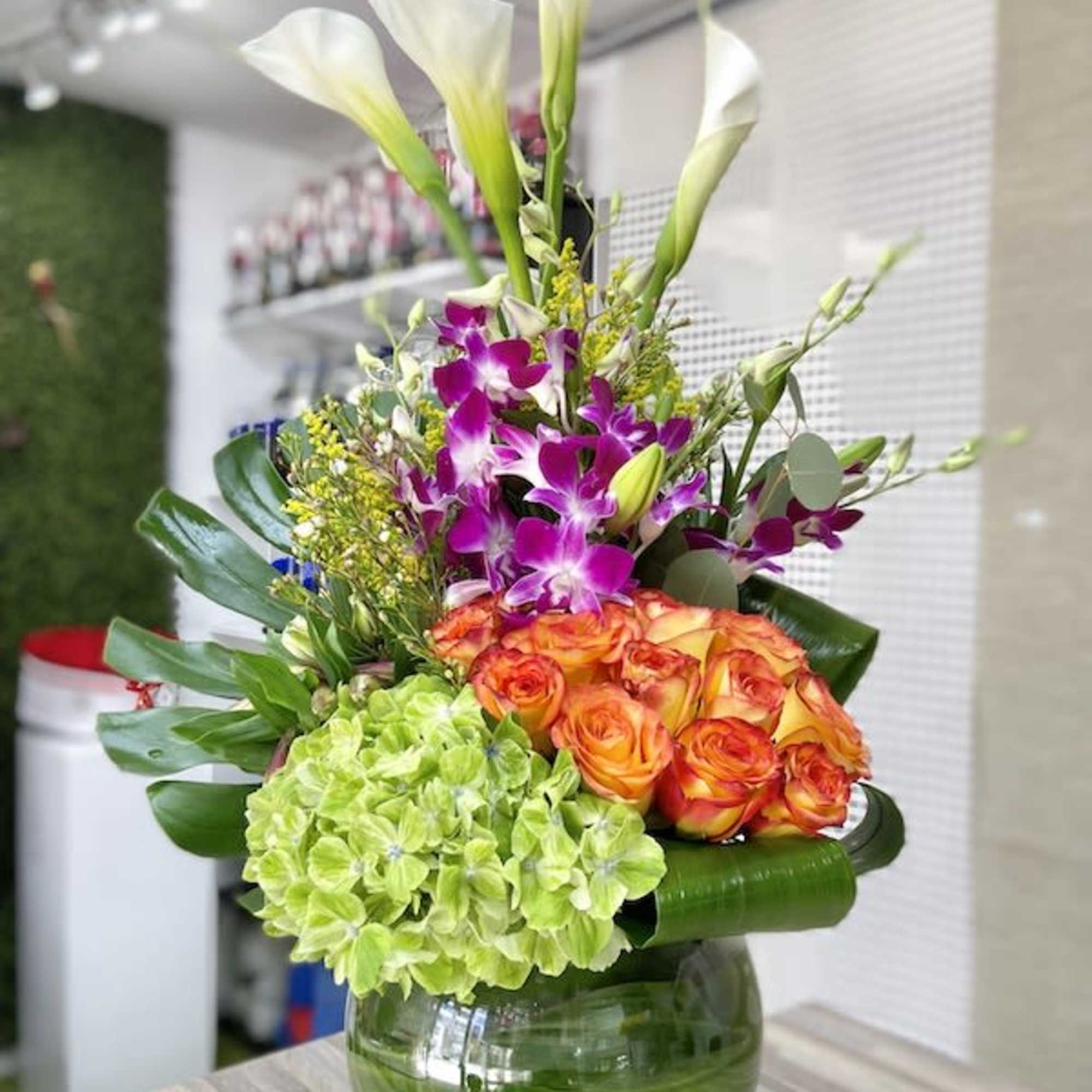 Tall mixed arrangement with calla lilies, orchids, roses, and hydrangea in a leaf-lined glass vase.