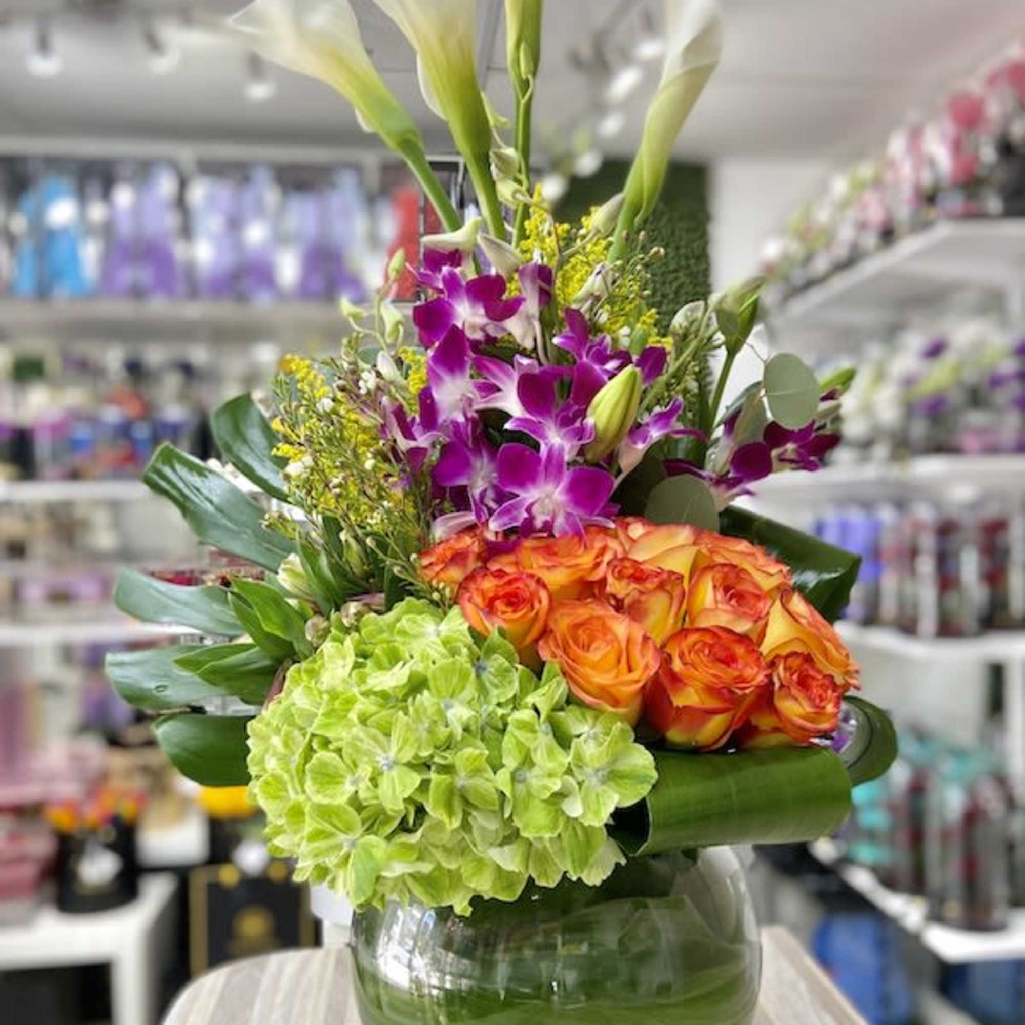 Tall arrangement with white calla lilies, purple orchids, orange roses, and green hydrangea in a round glass vase.