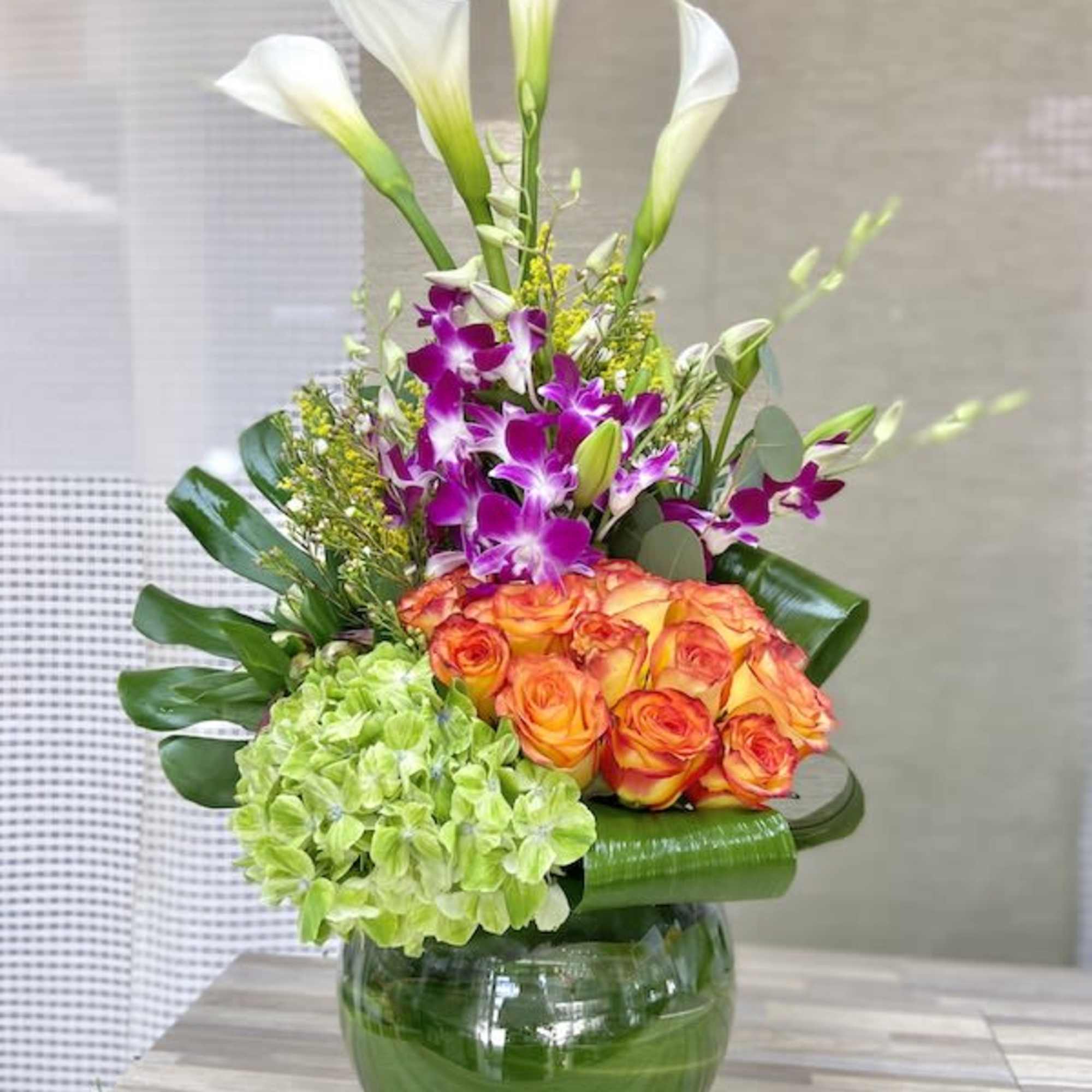 Tall arrangement of white calla lilies, purple orchids, orange roses, and green hydrangea in a round glass vase