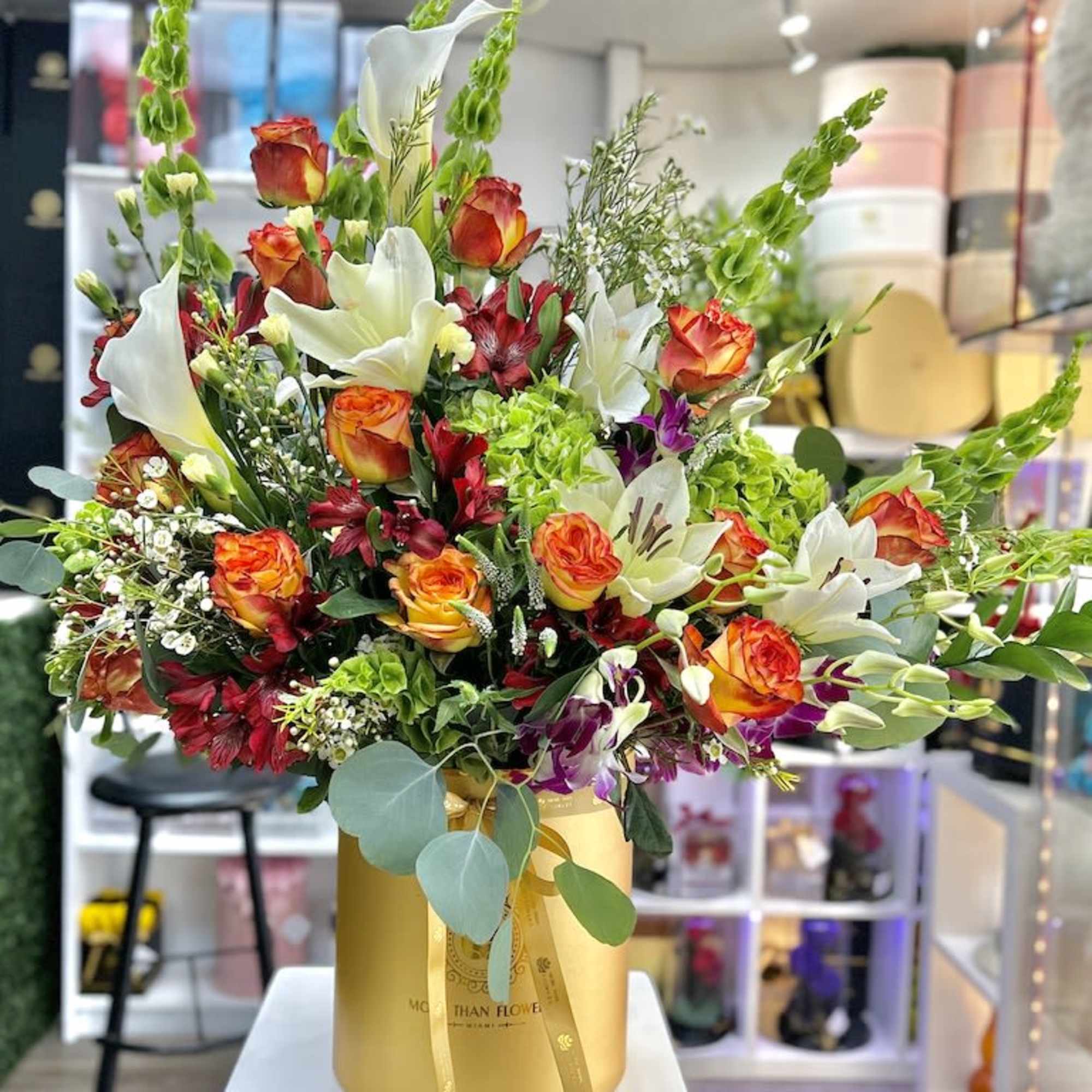Tall arrangement of orange roses, white lilies, and green accents in a gold hatbox container