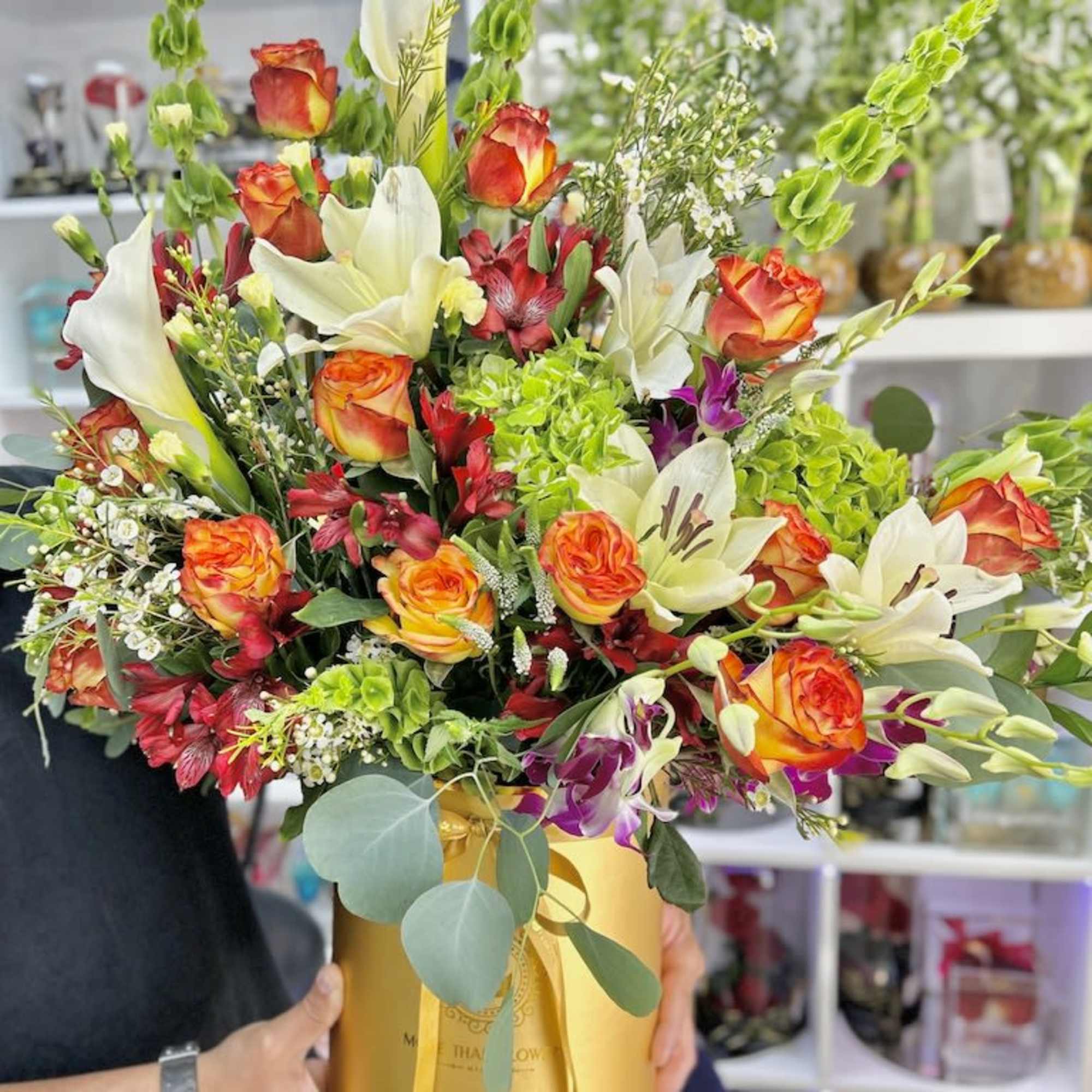 Tall arrangement of orange roses, white lilies, and green Bells of Ireland in a gold hatbox container