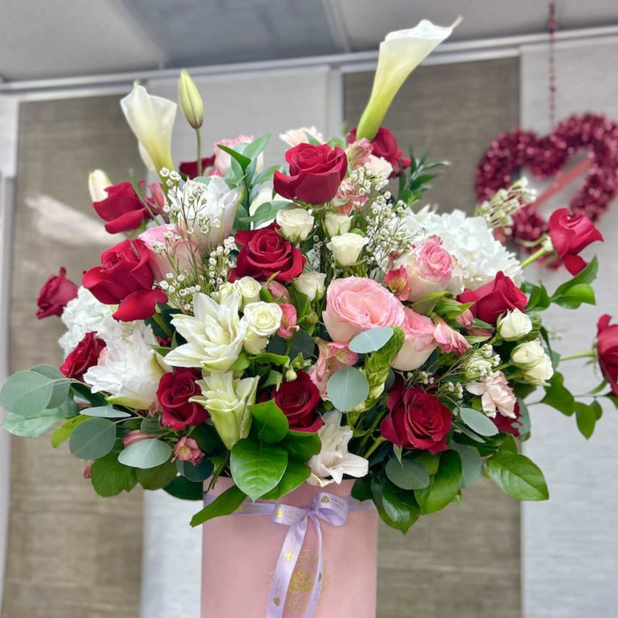 Tall arrangement of red and pink roses with white calla lilies in a pink hatbox-style container.
