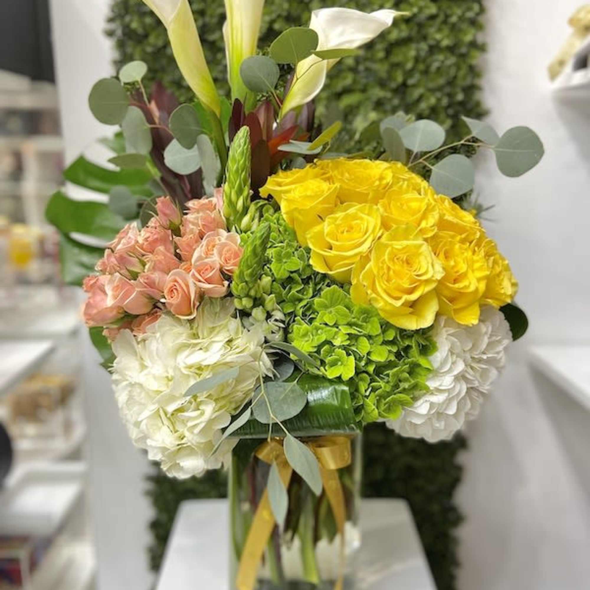 Tall arrangement of yellow roses, peach spray roses, white hydrangeas, and calla lilies in a glass vase