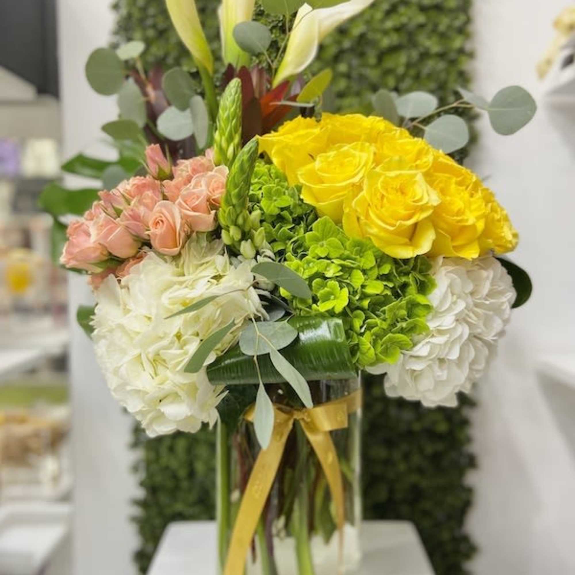 Tall glass vase with white calla lilies, yellow roses, peach spray roses, and hydrangeas.