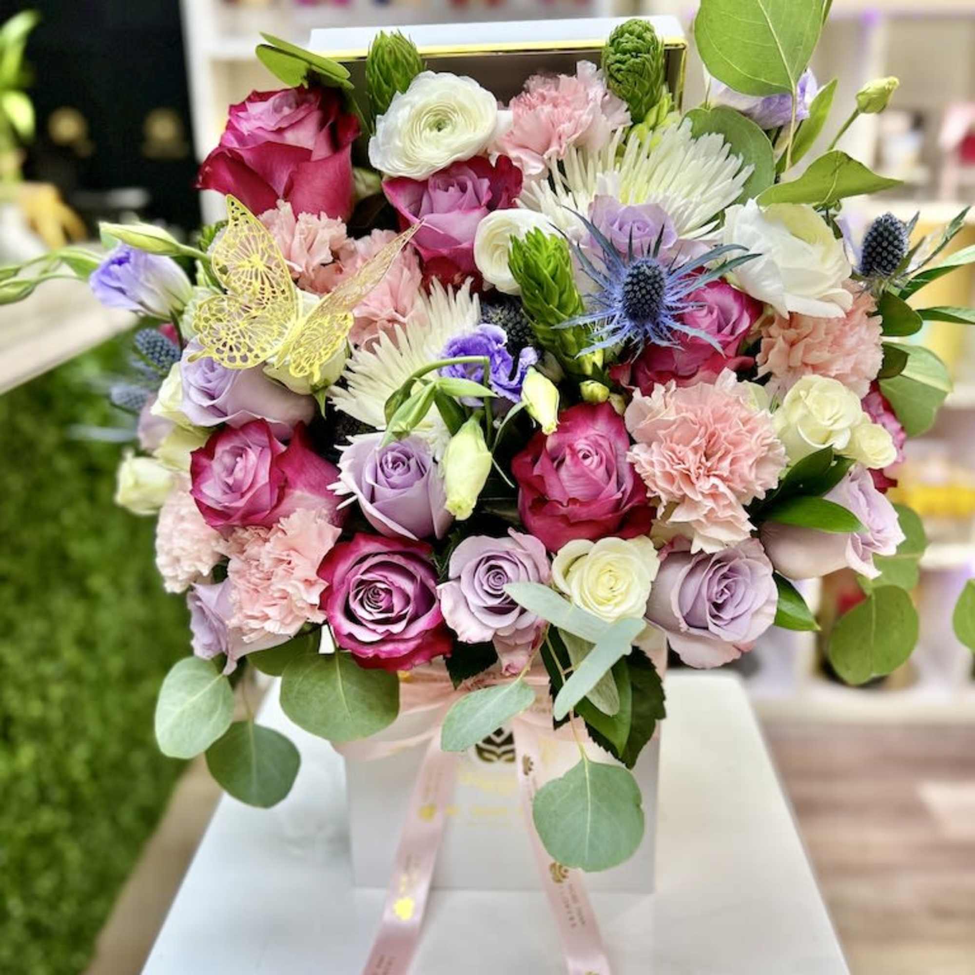 Box arrangement of pink, lavender, and white roses with mixed blooms and a butterfly pick