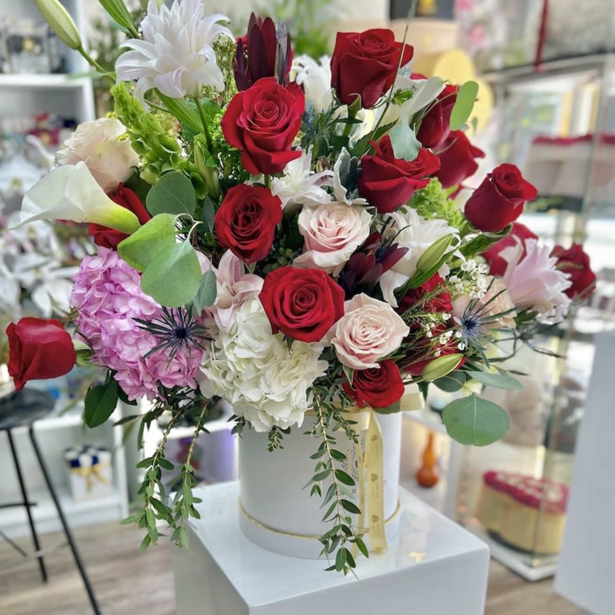 Tall mixed arrangement of red and blush roses, lilies, and hydrangeas in a white hatbox-style container.