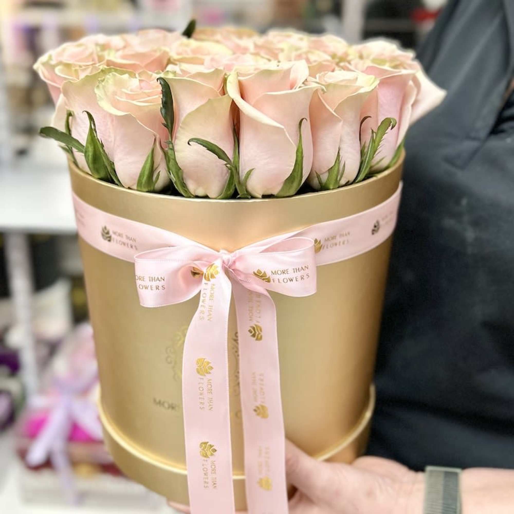 Blush pink roses arranged tightly in a round gold hatbox with a pink ribbon bow