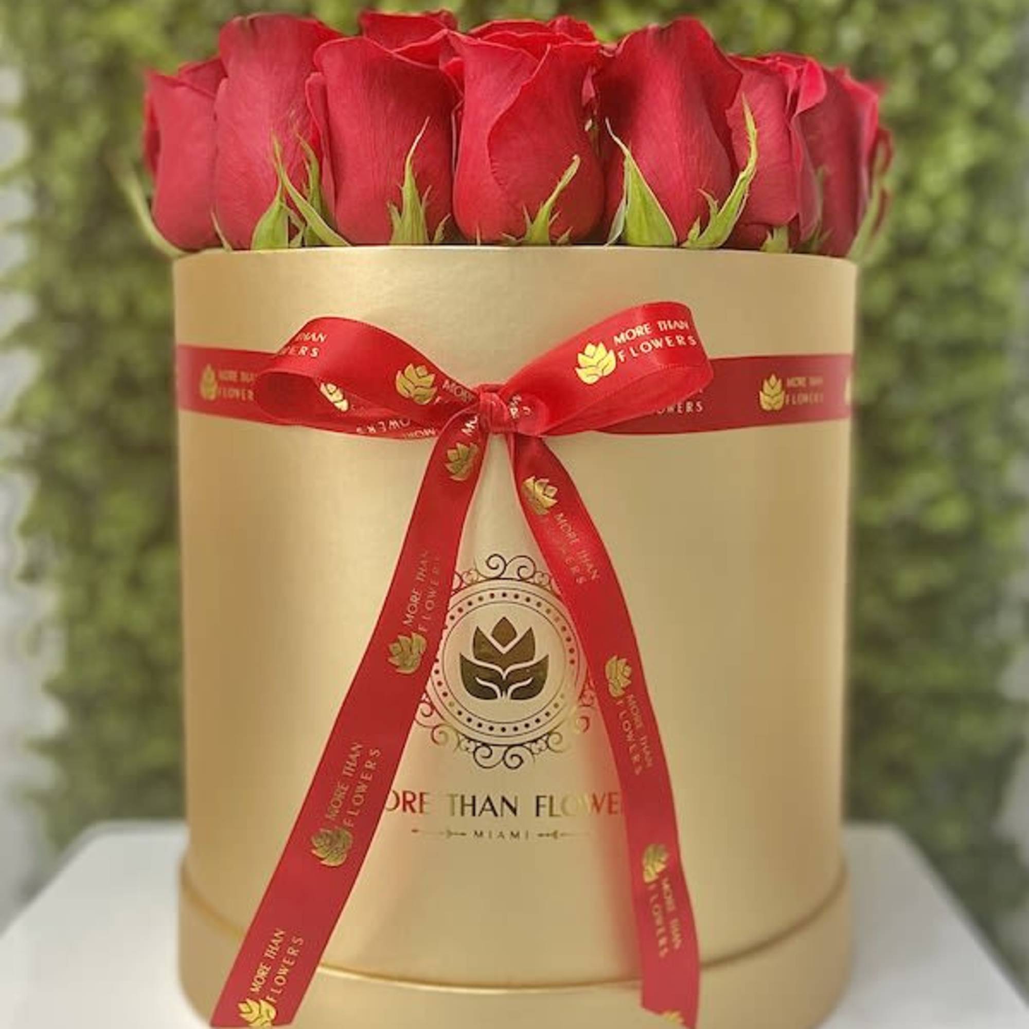 Round gold hat box filled with upright red roses and tied with a red ribbon.
