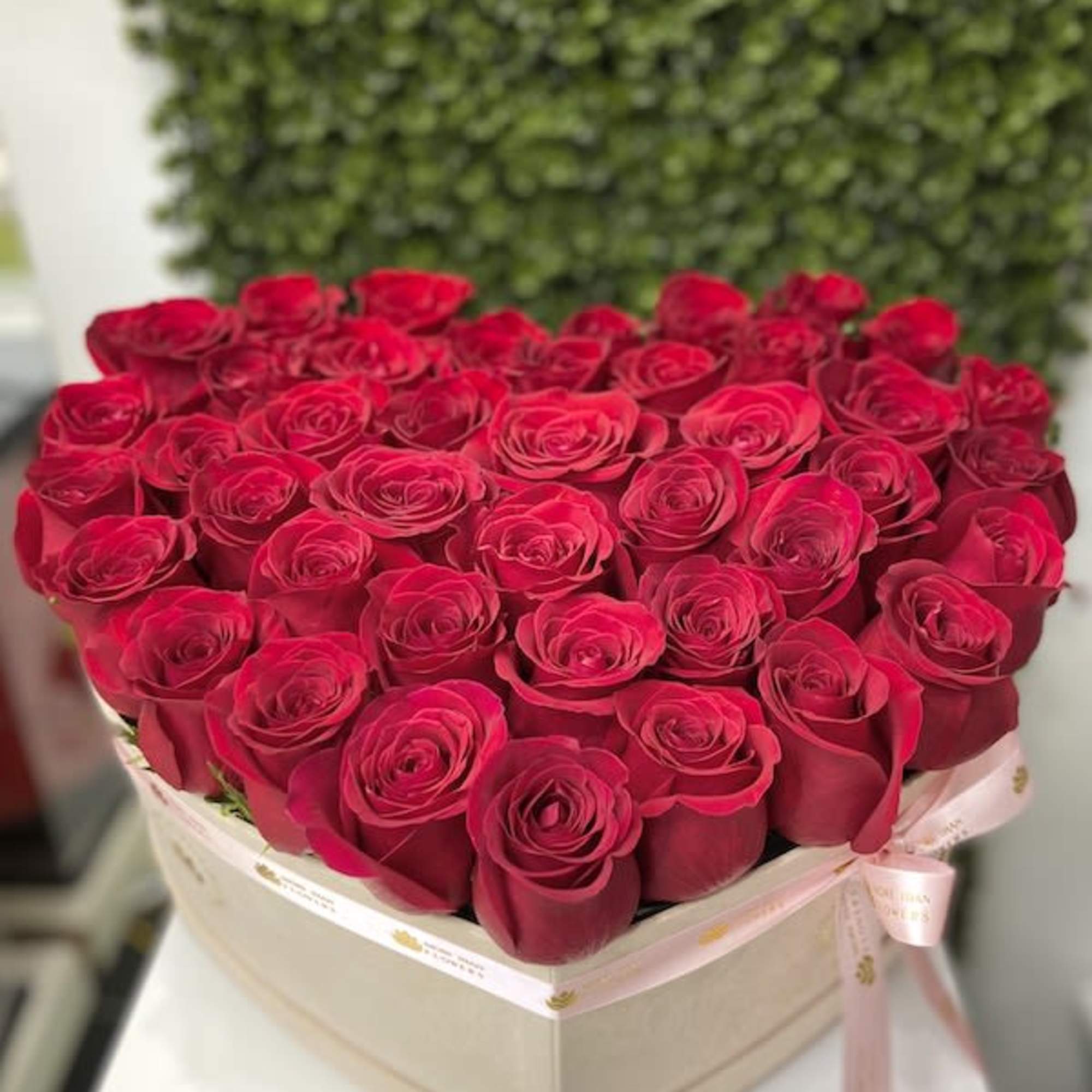 Heart-shaped box filled with tightly packed red roses and tied with a pink ribbon.