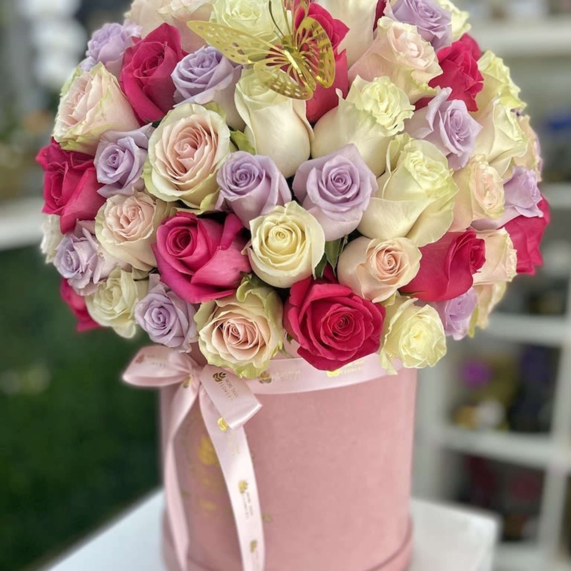 Round hatbox of pink, white, cream, and lavender roses with a gold butterfly accent and ribbon bow.