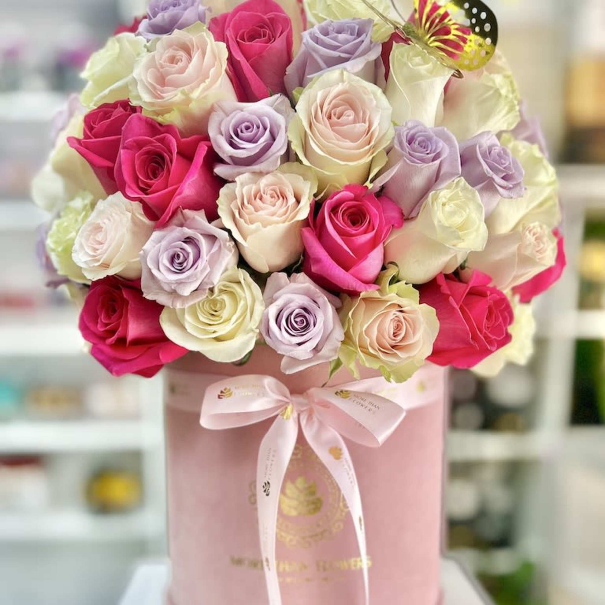 Round pink hatbox filled with pink, white, and lavender roses with a ribbon bow and butterfly accent.