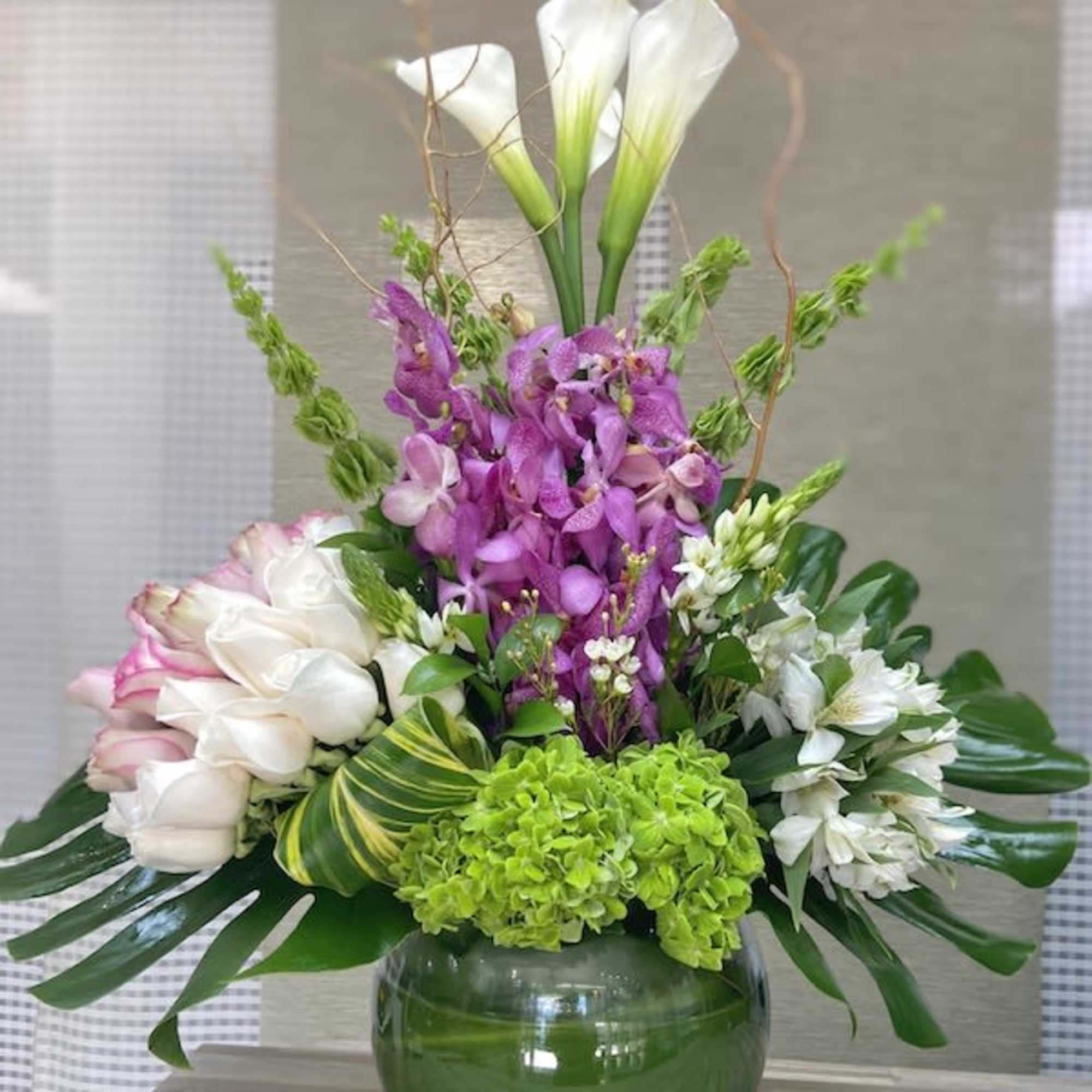 Tall arrangement with white calla lilies, purple orchids, green hydrangeas, and roses in a round green glass vase