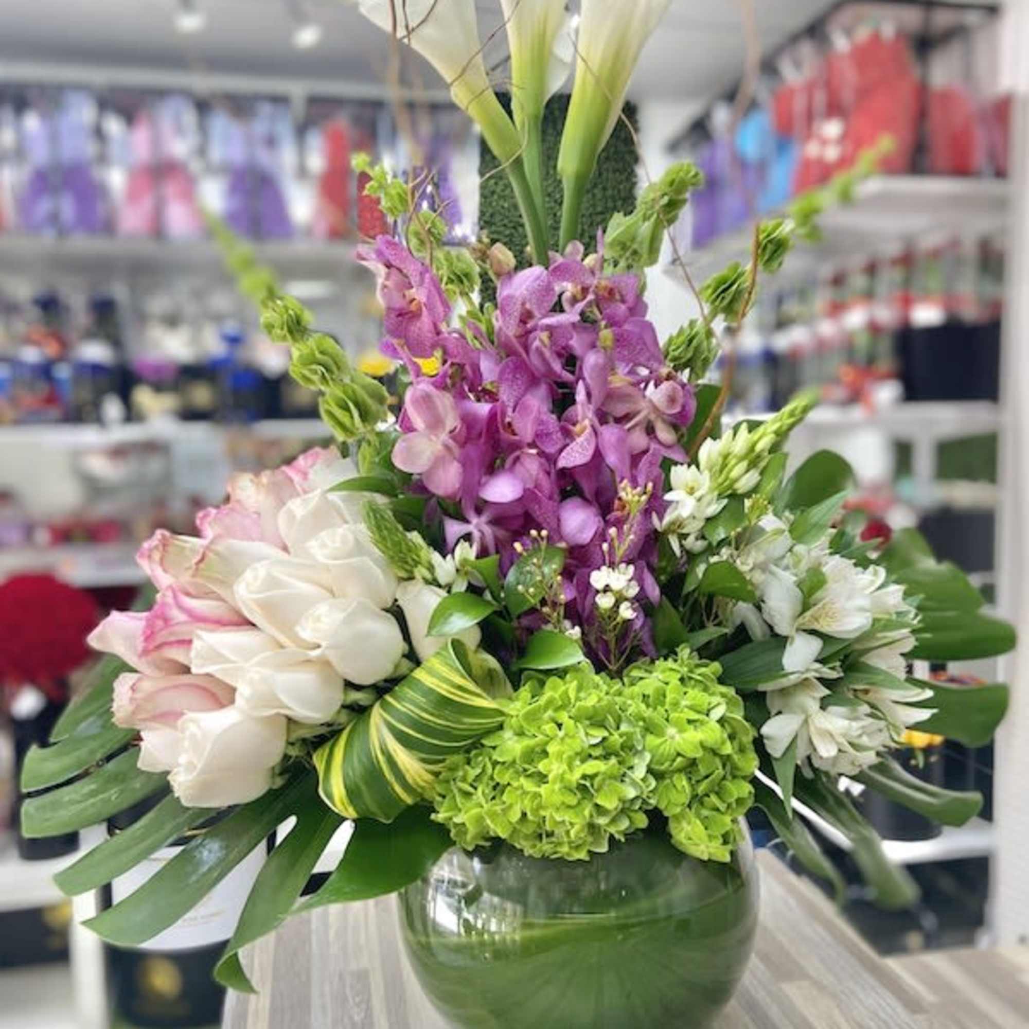 Tall mixed arrangement with white calla lilies, purple orchids, and green hydrangeas in a round glass vase
