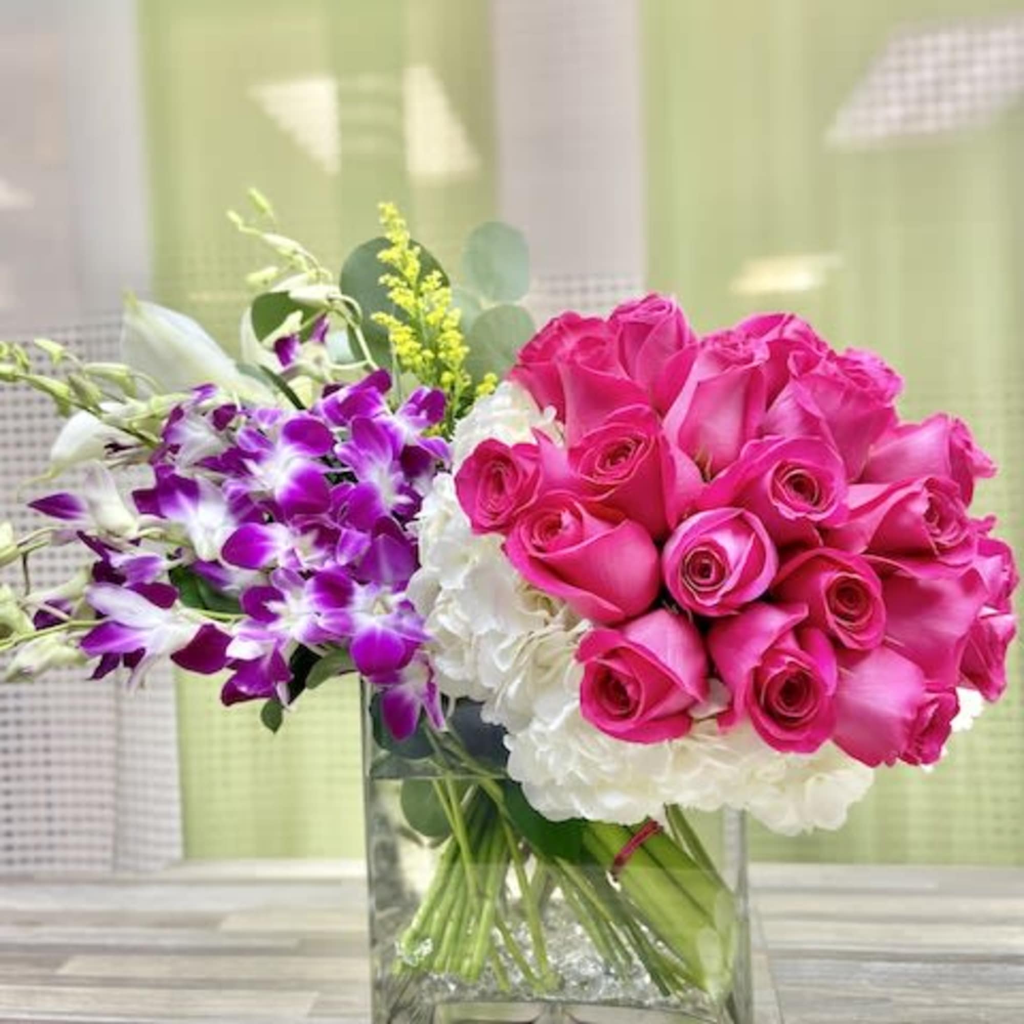 Modern arrangement of hot pink roses, white hydrangea, and purple orchids in a clear square glass vase