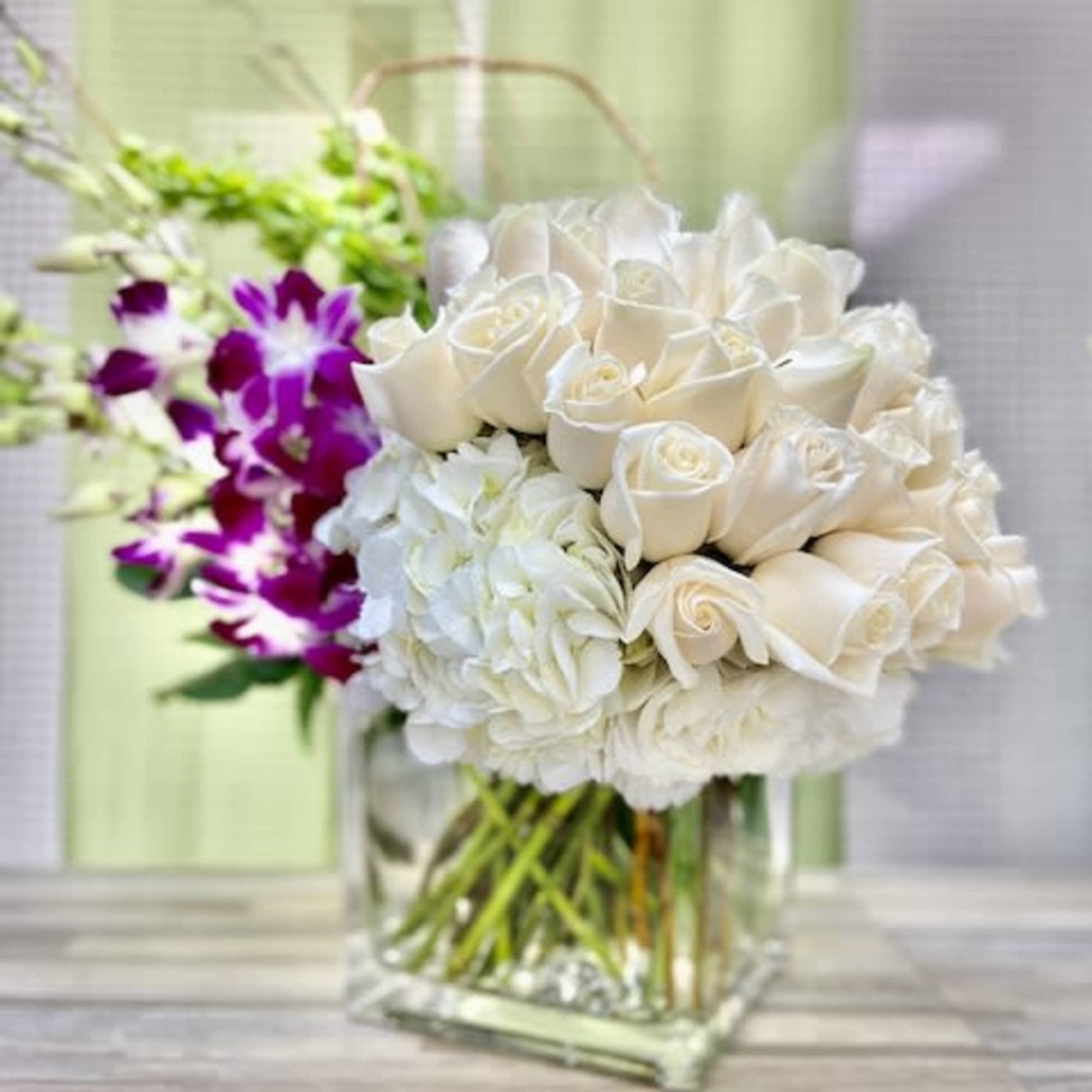 White roses and hydrangeas in a clear cube vase with purple orchids and green Bells of Ireland in the background.