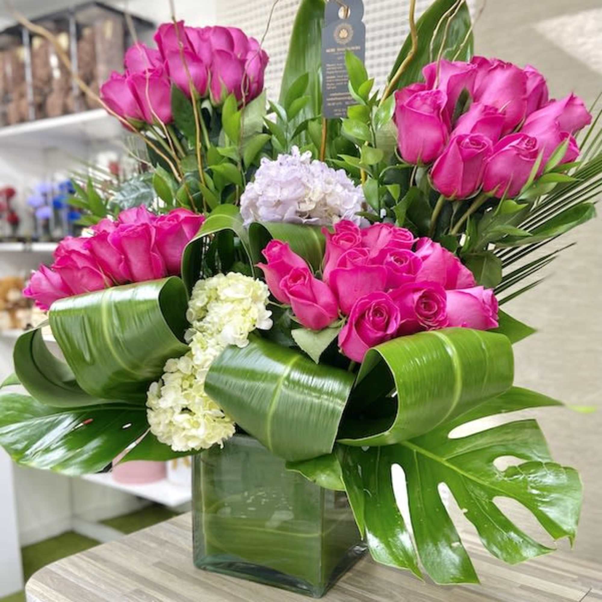 Tall arrangement of hot pink roses with white hydrangeas and large green tropical leaves in a glass cube vase