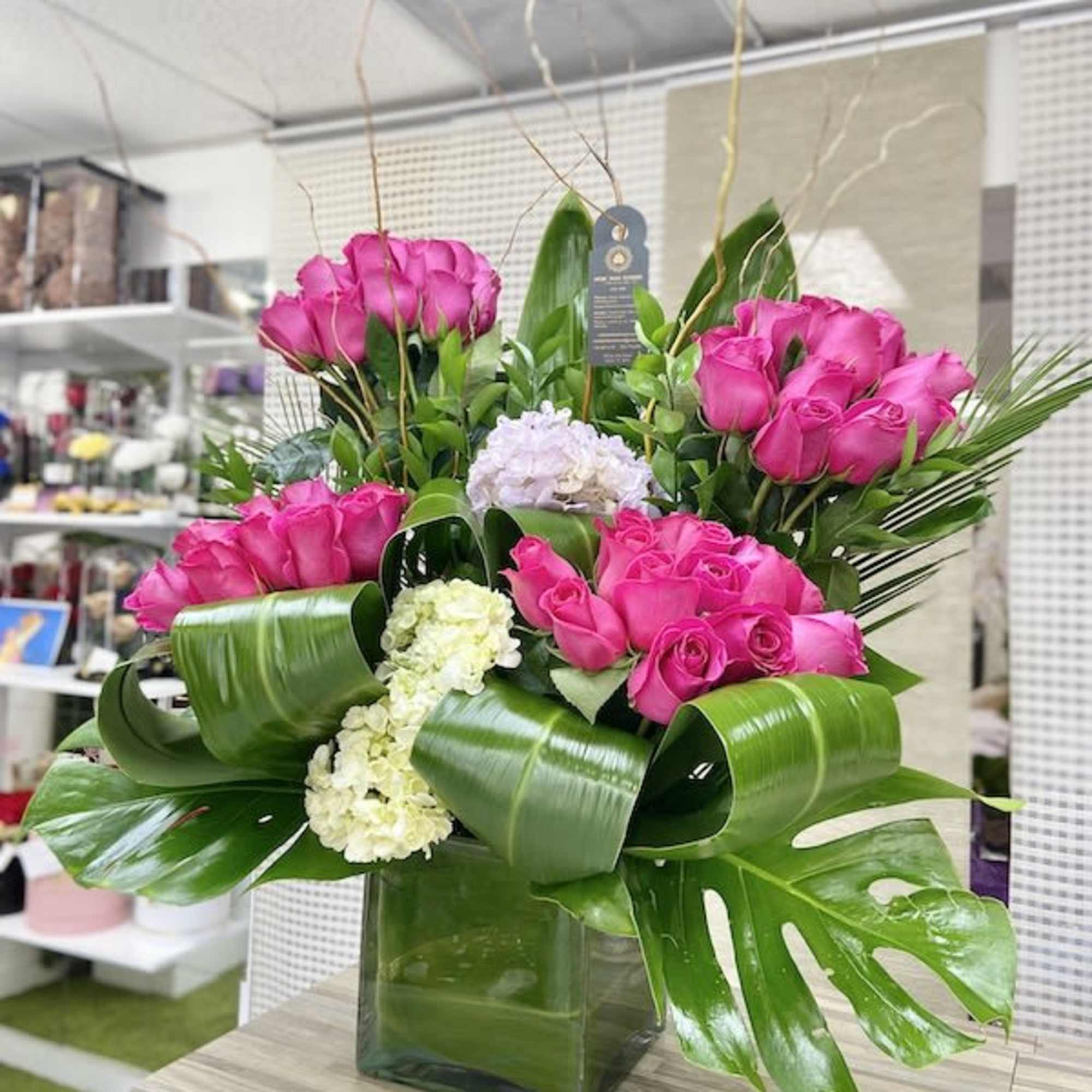 Tall arrangement of hot pink roses and white hydrangeas with large green leaves in a square glass vase