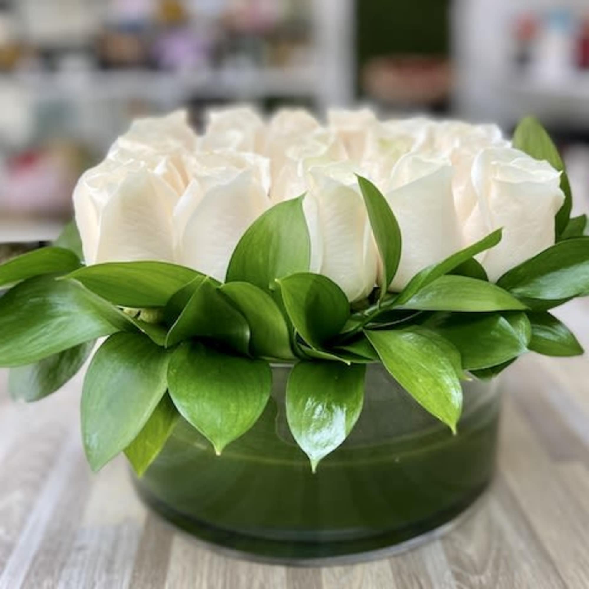 Low arrangement of white roses in a round glass vase bordered with glossy green leaves