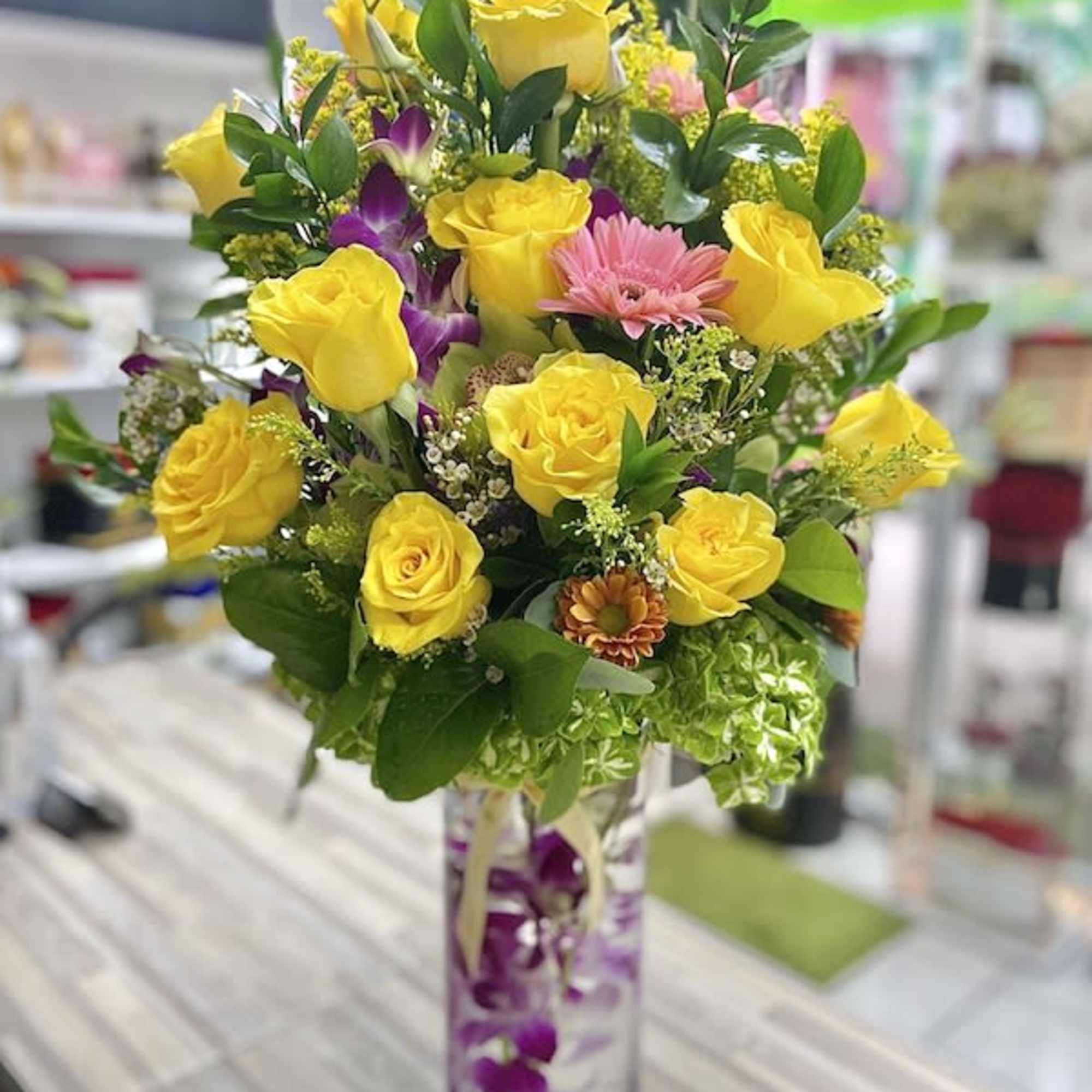 Tall arrangement of yellow roses, pink daisies, and orchids in a clear cylinder vase