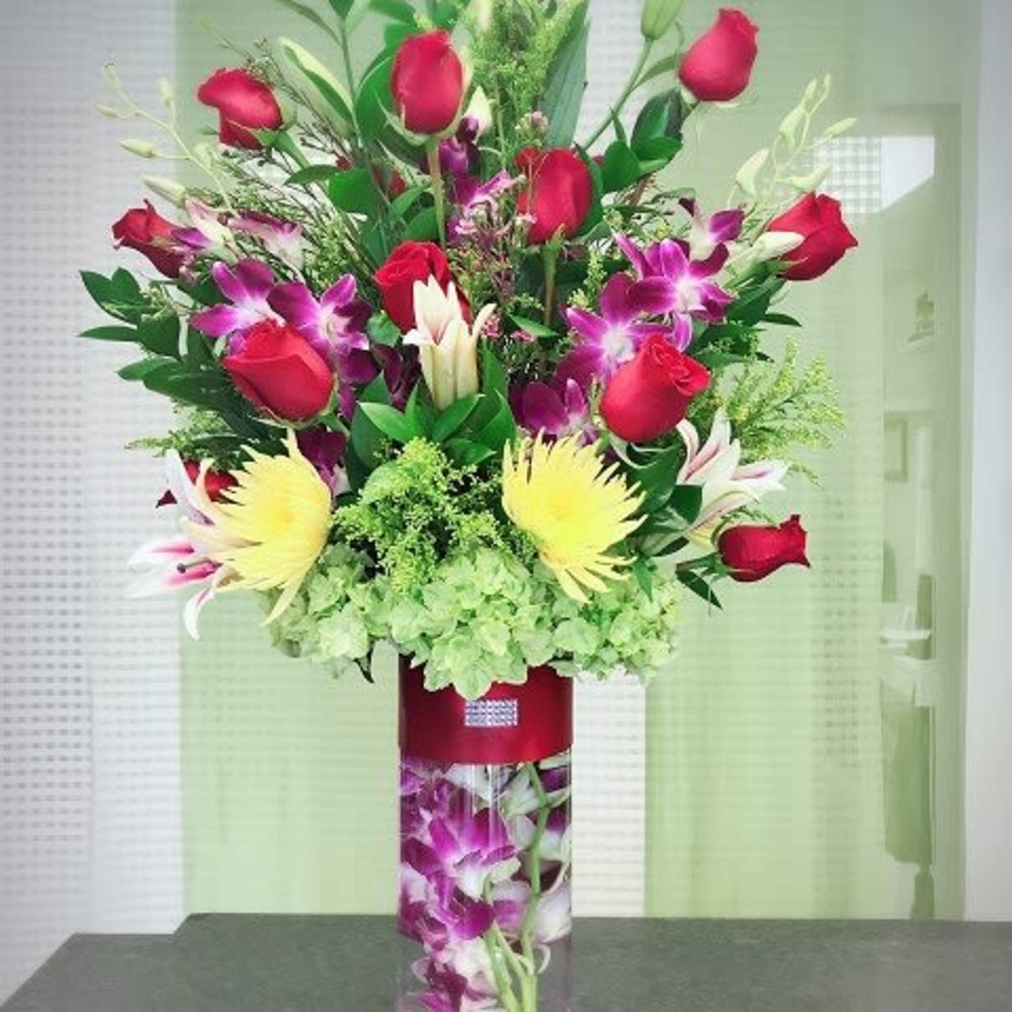 Tall arrangement of red roses, purple orchids, lilies and yellow blooms in a clear cylinder vase