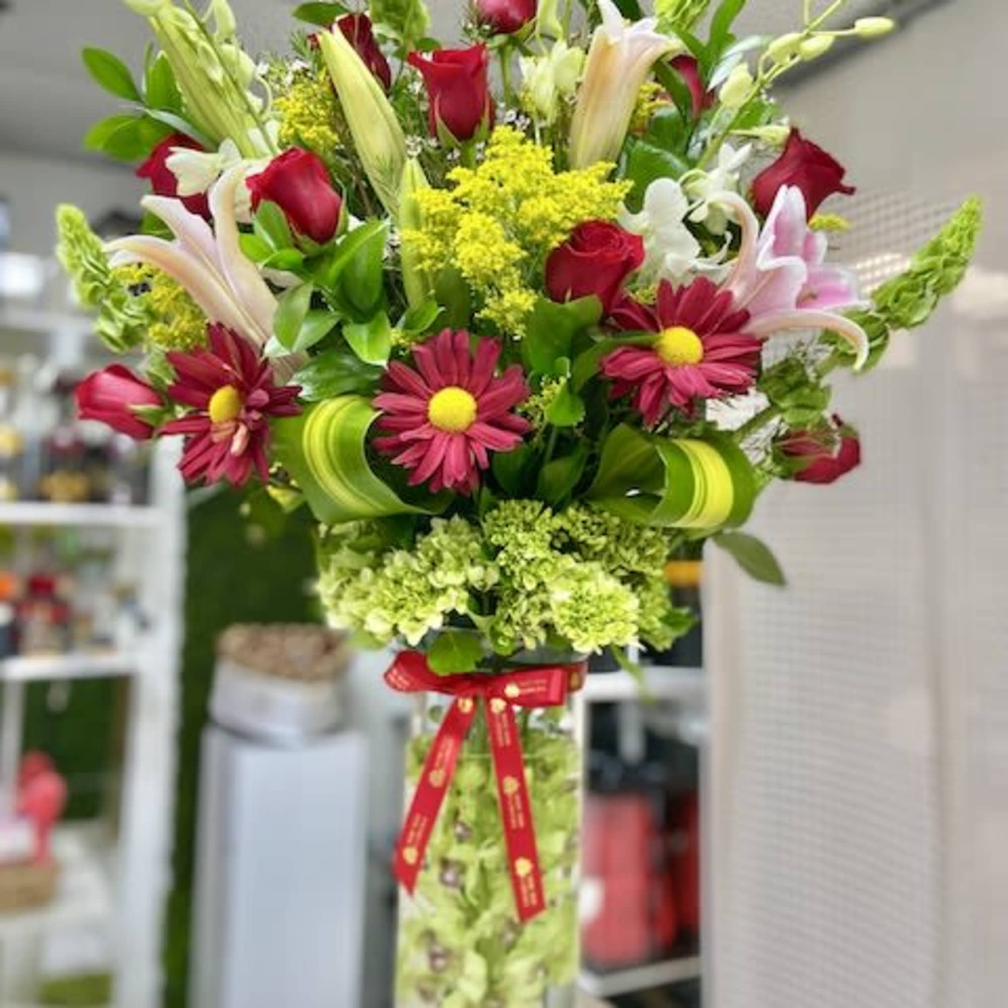 Tall mixed arrangement of red roses, pink lilies, and daisies in a clear cylinder vase with a red ribbon