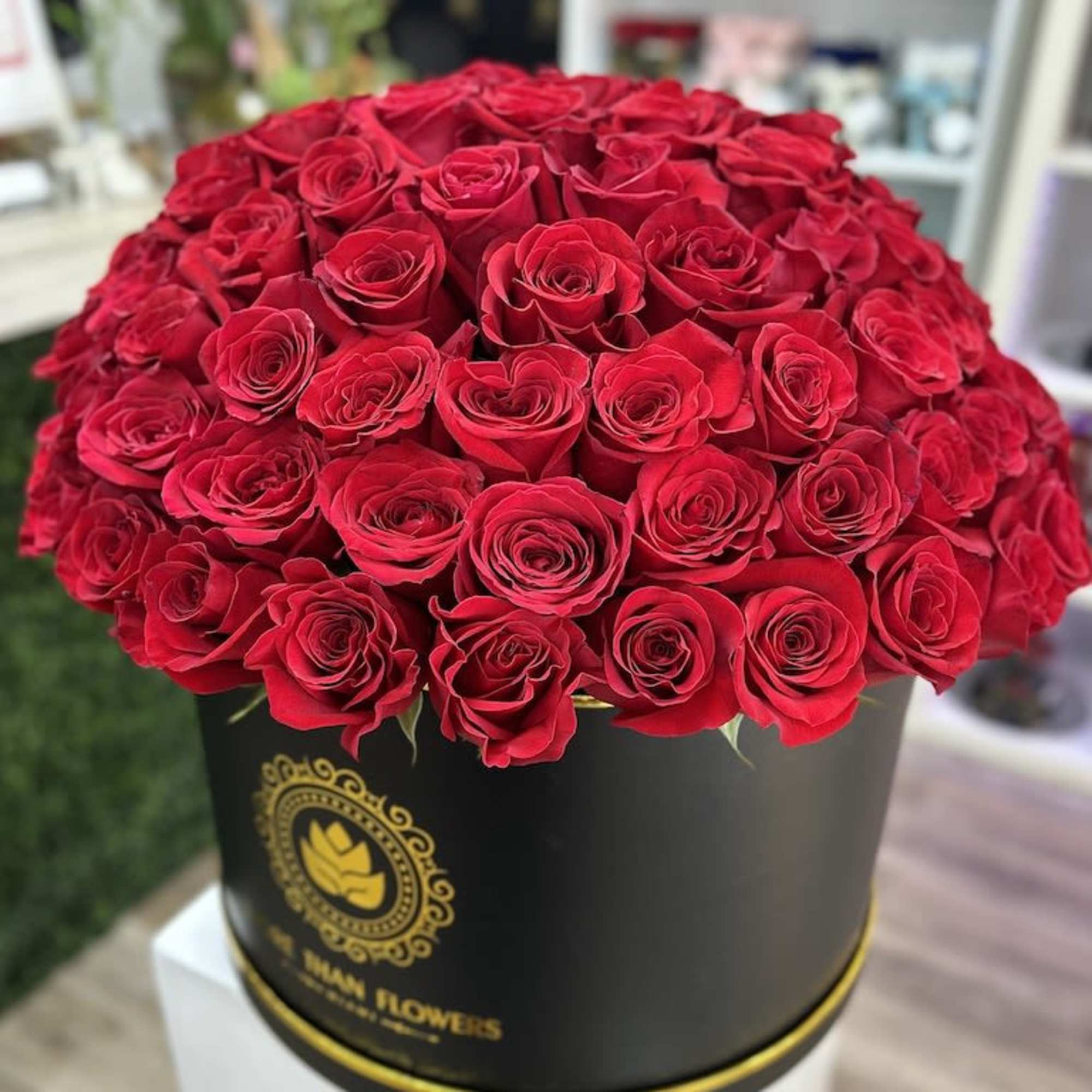 Round black hatbox filled with many red roses on a white pedestal.