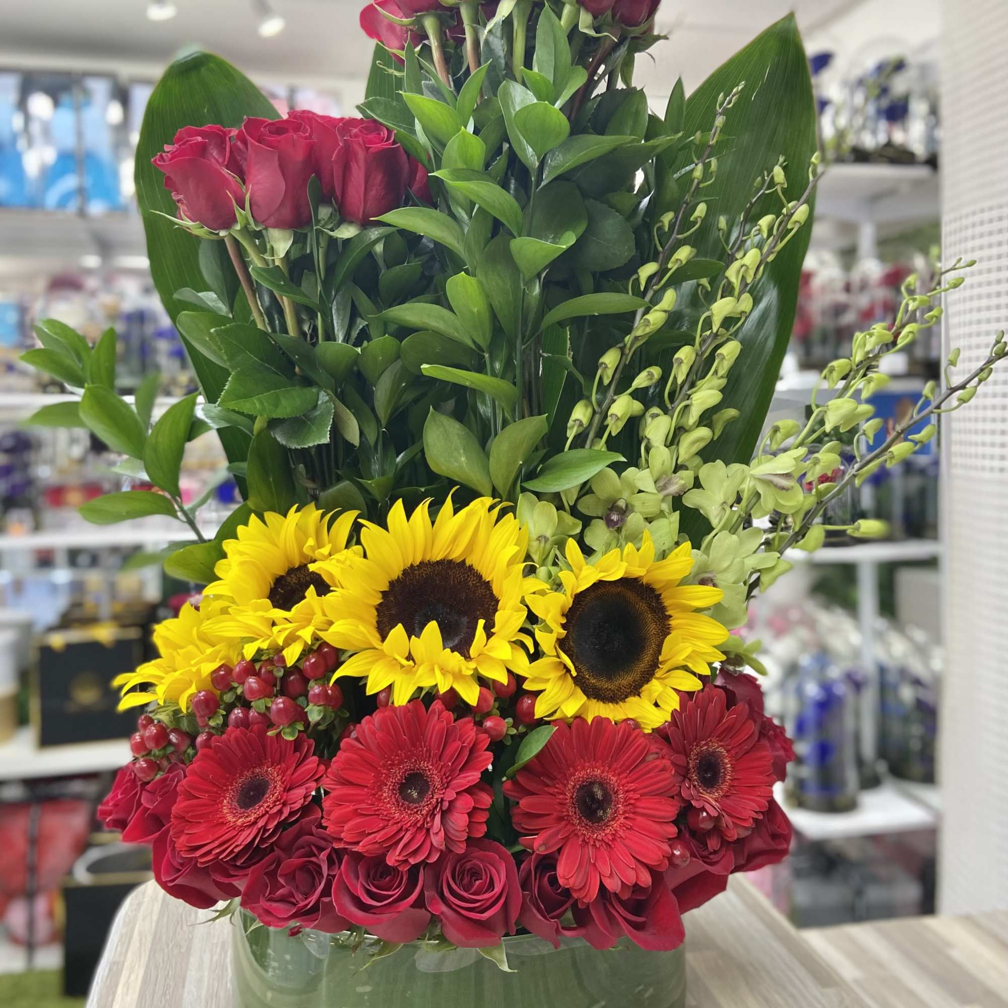 Tall arrangement of red roses, red gerbera daisies, and yellow sunflowers in a round glass vase