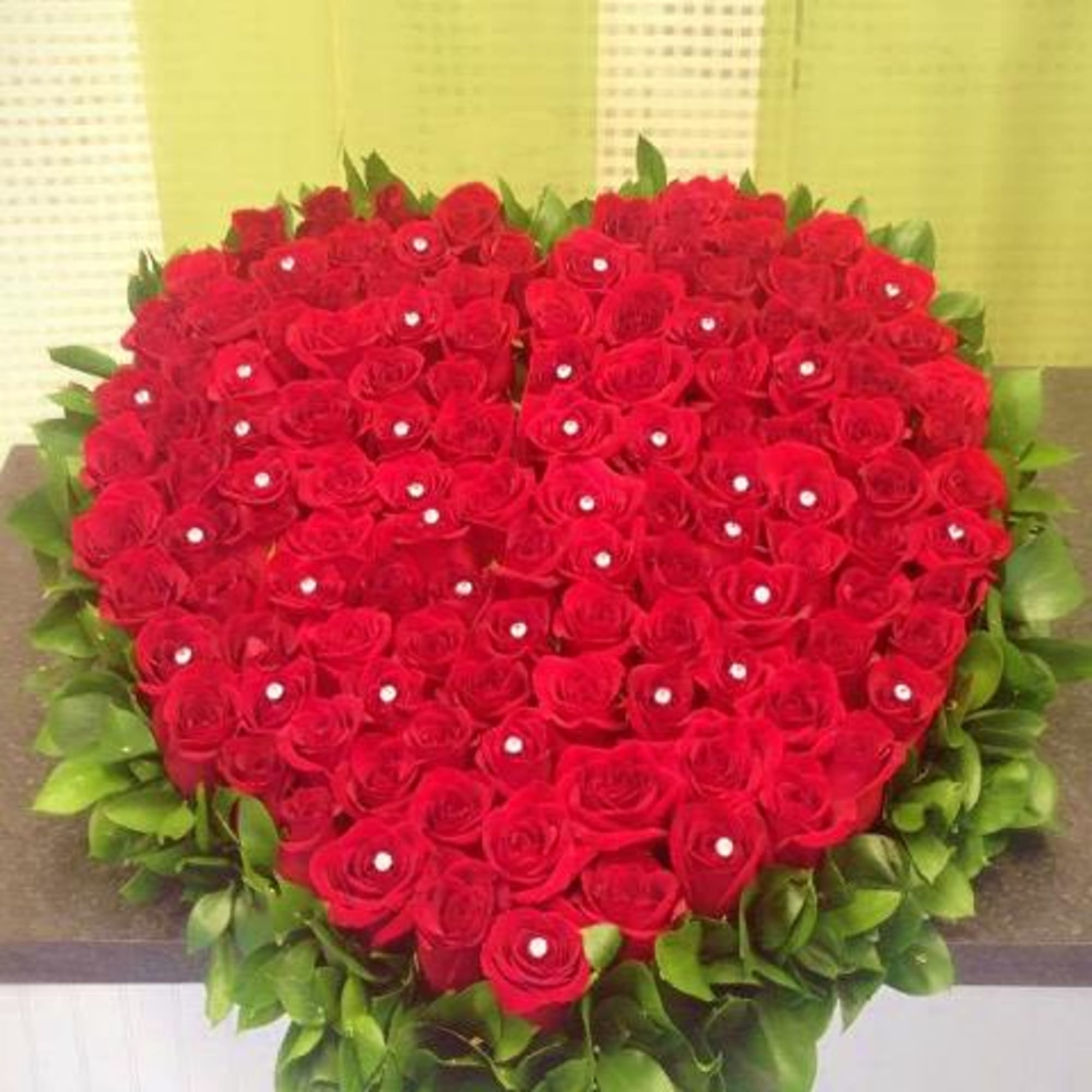 Heart-shaped arrangement of red roses with jeweled centers bordered by glossy green foliage
