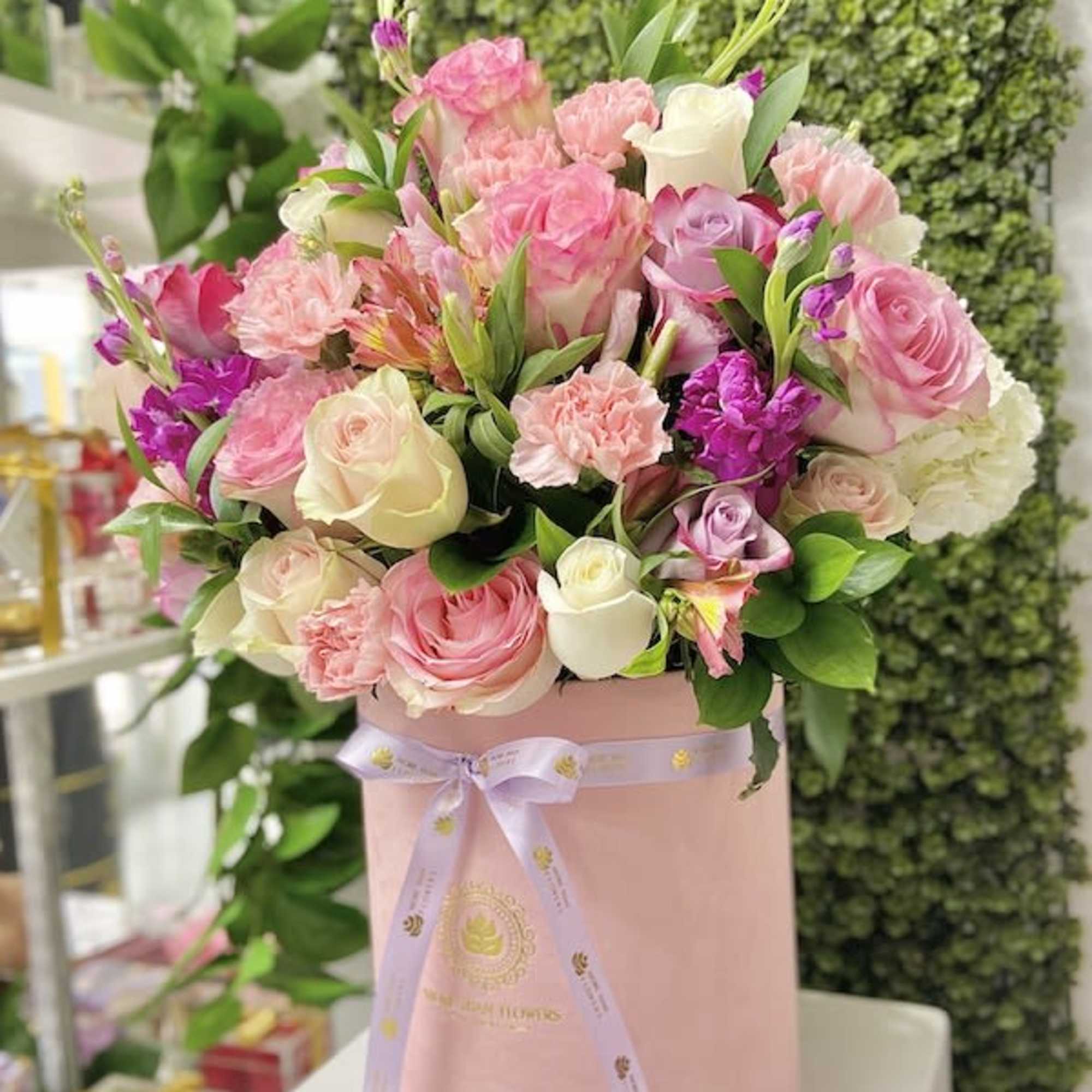 Round pink hatbox filled with pink, white, and purple mixed roses and carnations.