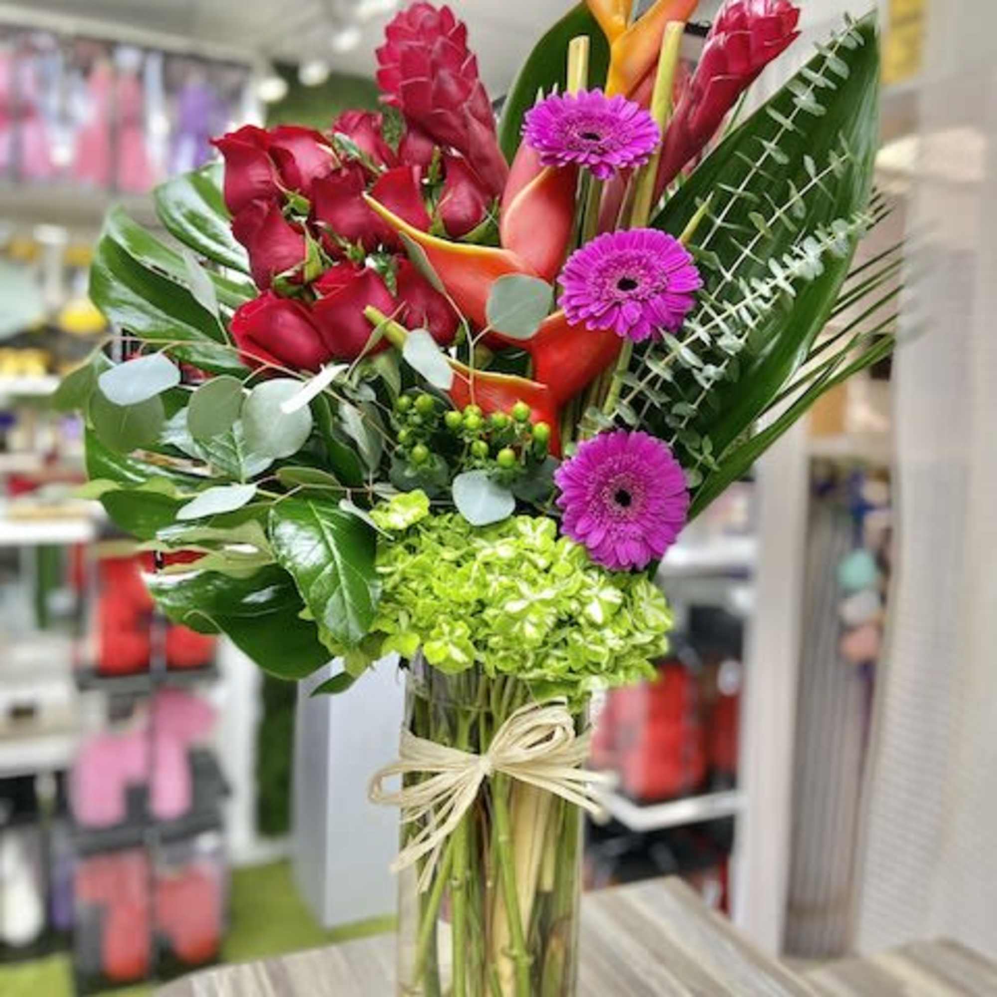 Tall tropical arrangement with red roses, orange blooms, green hydrangea and purple daisies in a glass vase