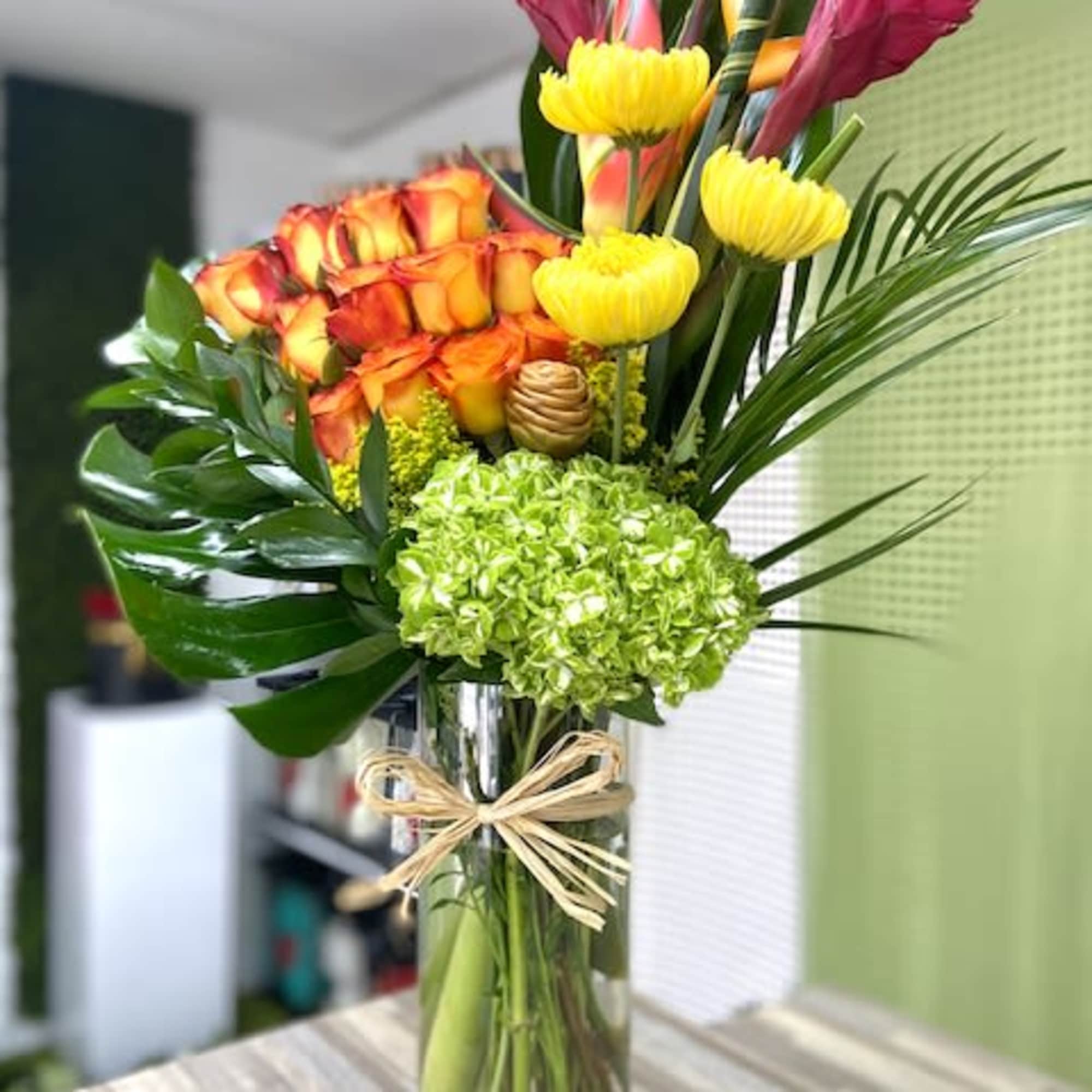 Tall tropical arrangement with orange roses, green hydrangea and yellow blooms in a glass vase with raffia bow