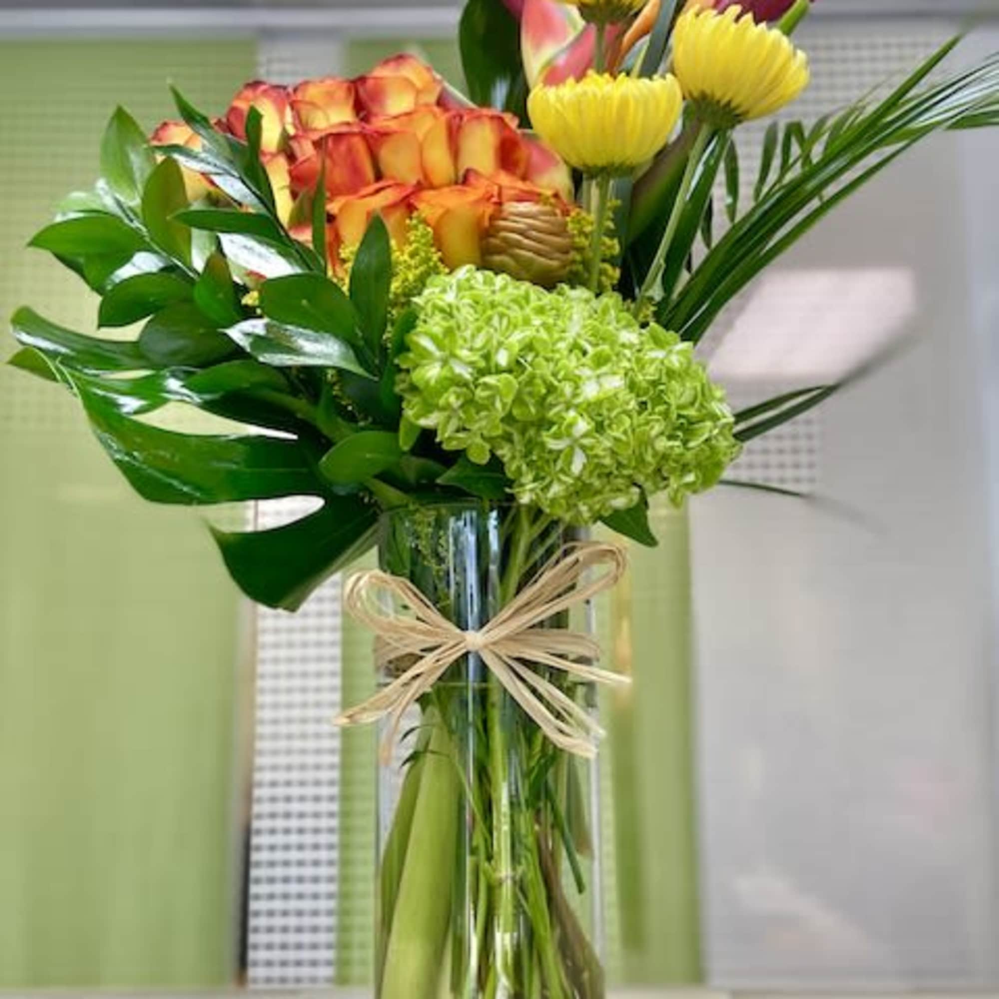 Tall mixed arrangement of orange roses, yellow mums, green hydrangea and tropical flowers in a glass vase