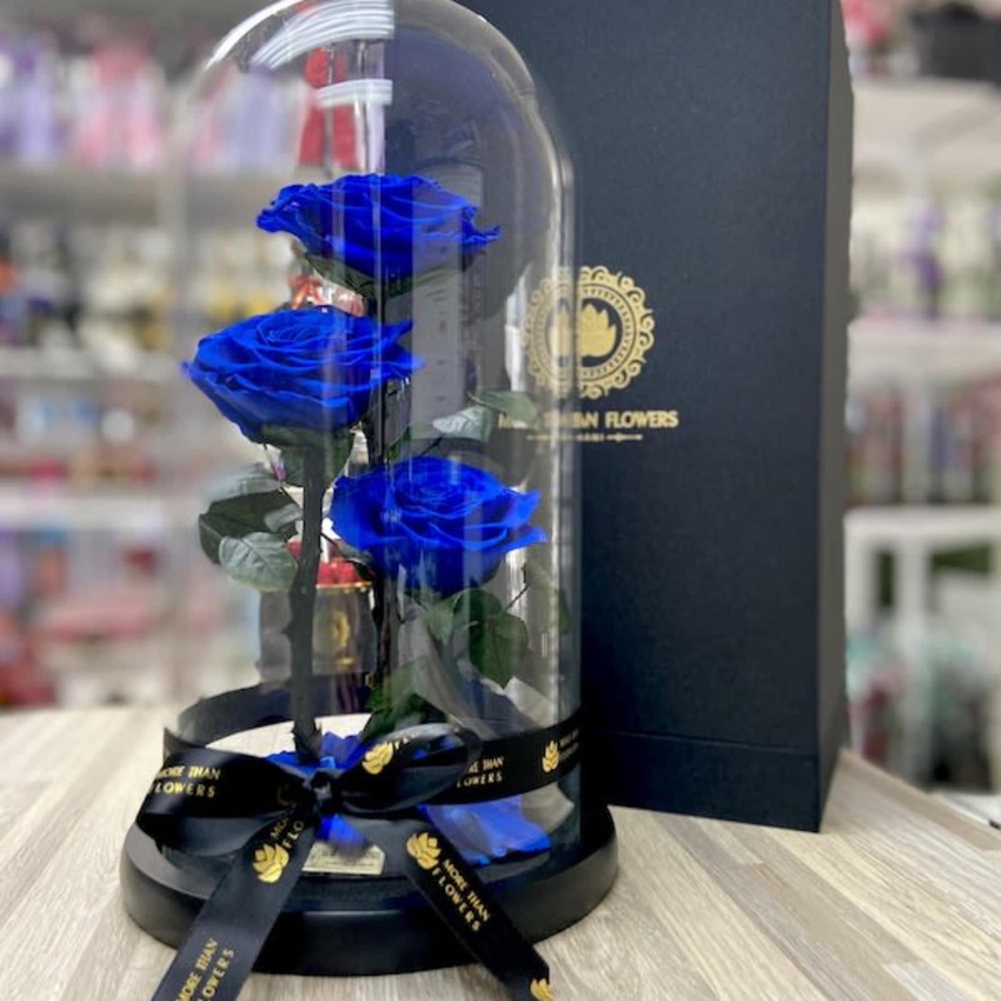 Three blue roses under a clear glass dome on a black base with ribbon and gift box.