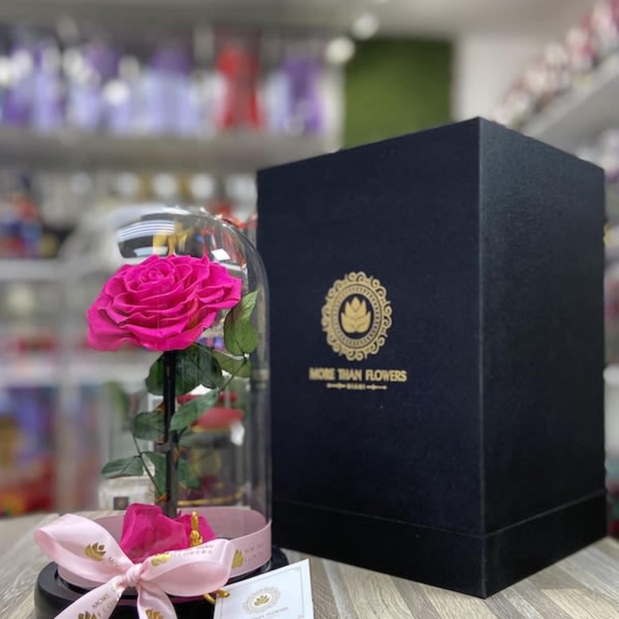 Single bright pink rose under a glass dome on a black base with a tall black gift box beside it.