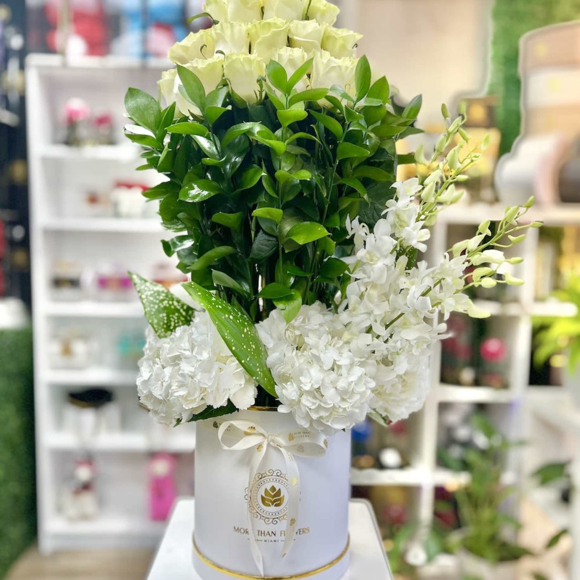 Tall white hatbox arrangement with cream roses, white hydrangeas, orchids, and green foliage.