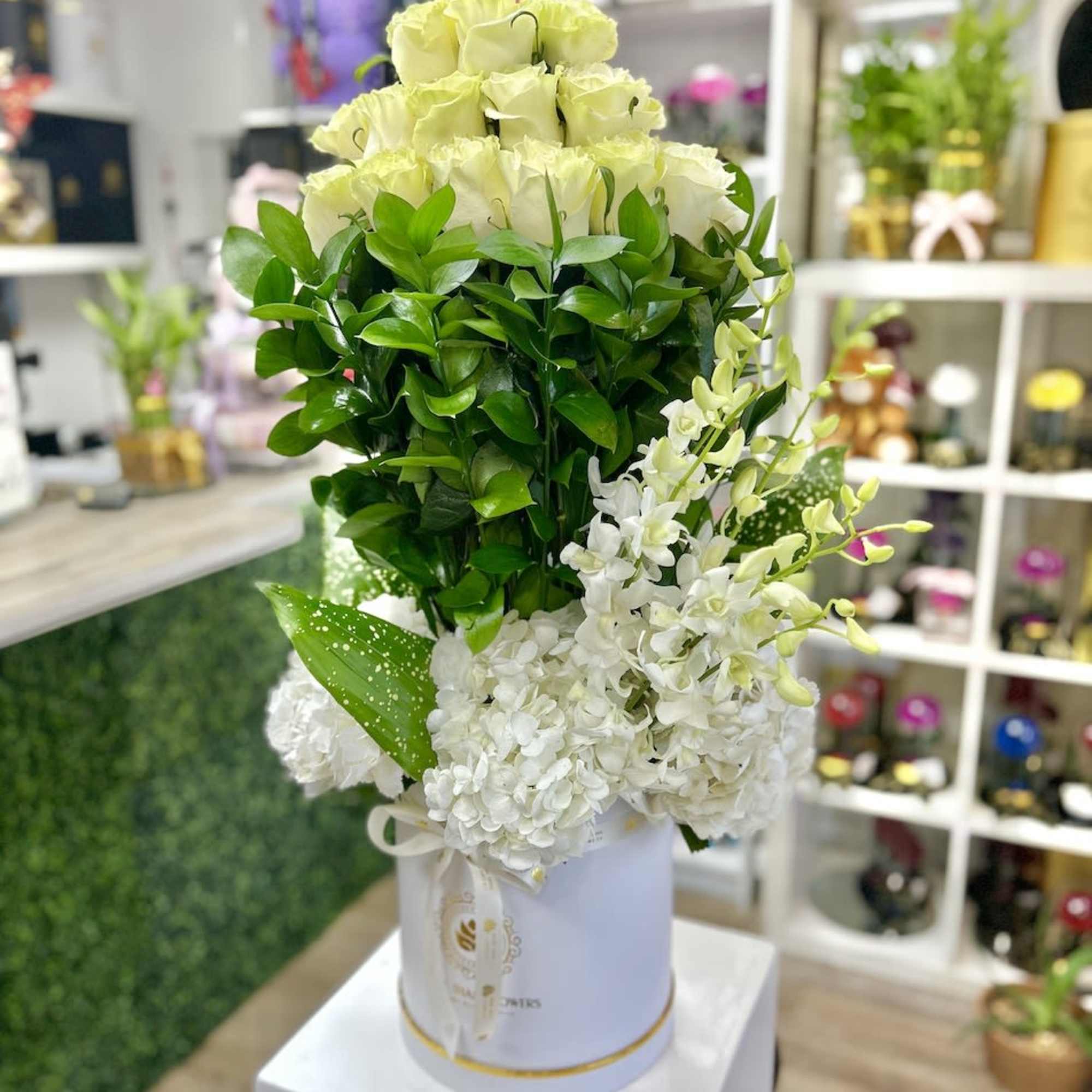 Tall arrangement of pale roses, white blooms, and dense greenery in a white hatbox container